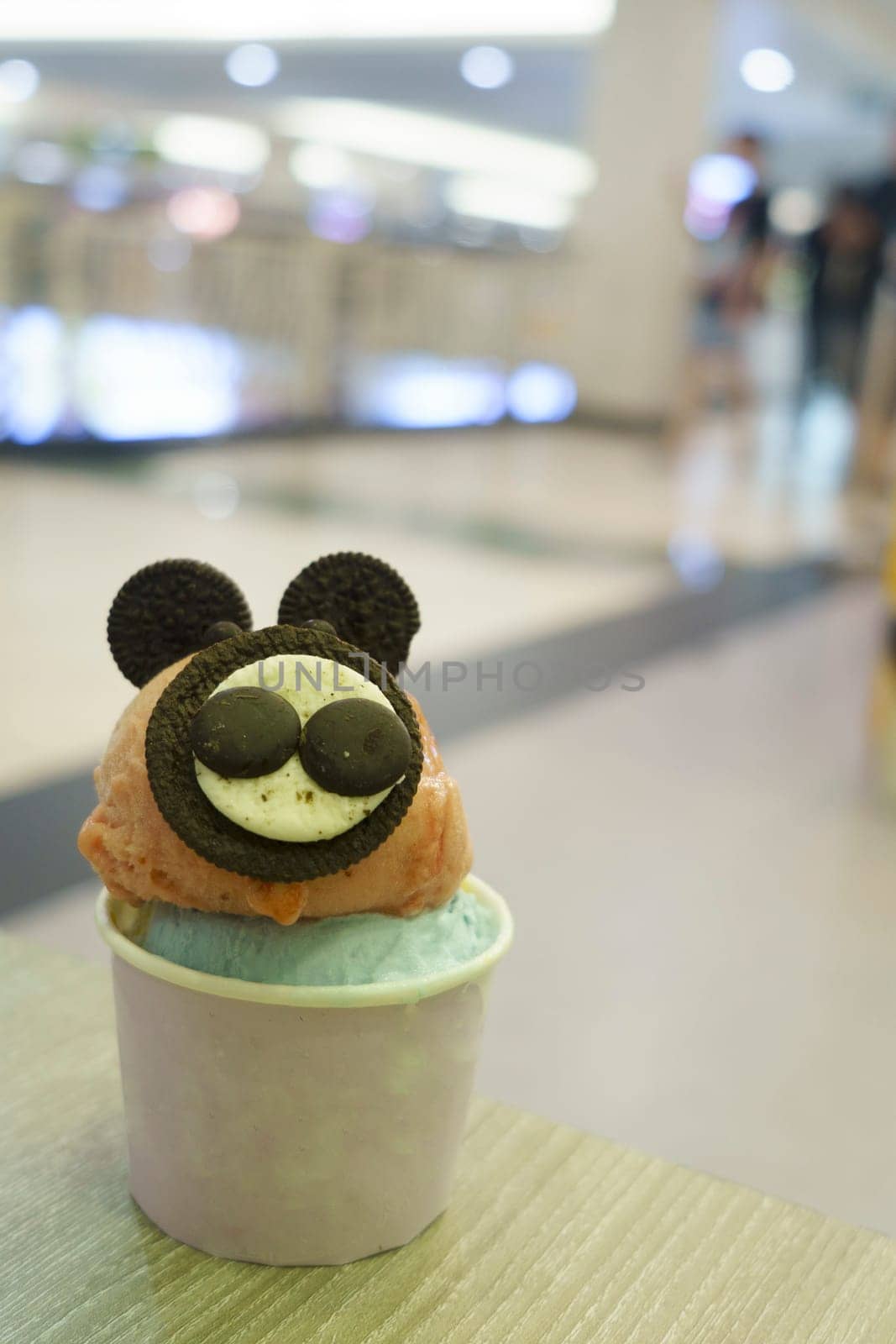 A cup of gelato with mouse-like biscuit topping served on the wooden table with blurred background.