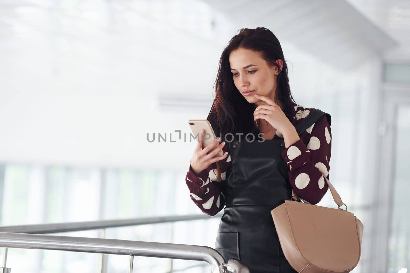 Holding phone. Beautiful young brunette in black skirt indoors in office or airport. Having free time.