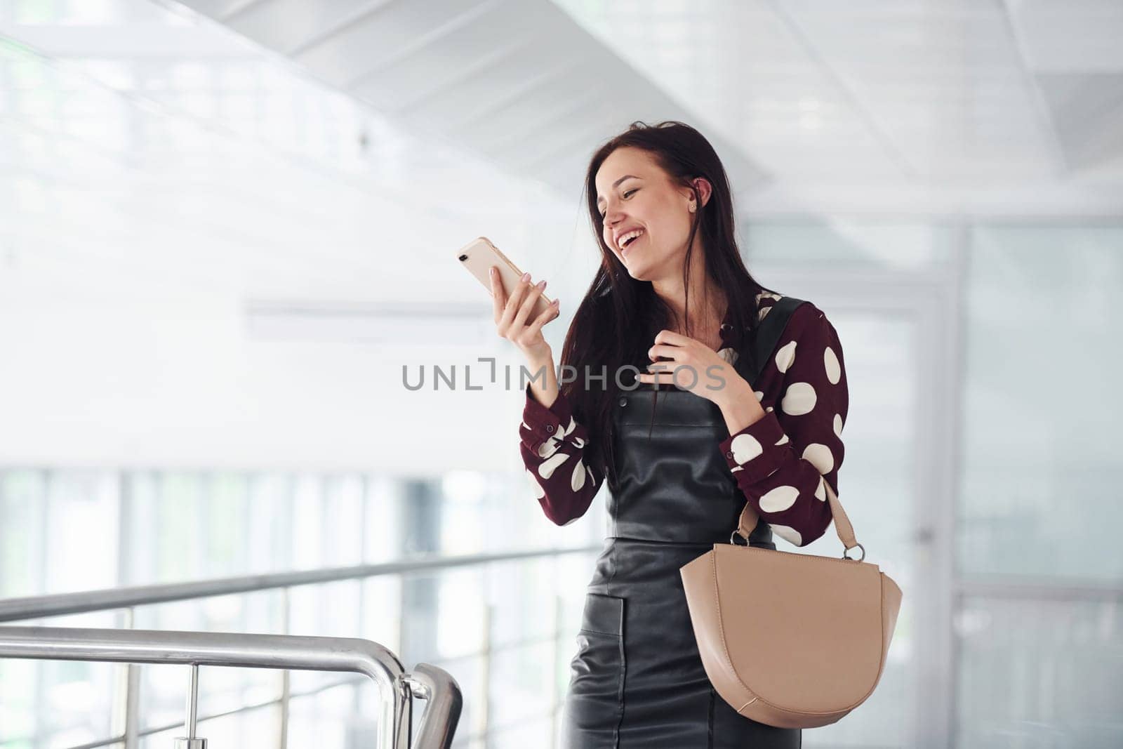 Holding phone. Beautiful young brunette in black skirt indoors in office or airport. Having free time by Standret