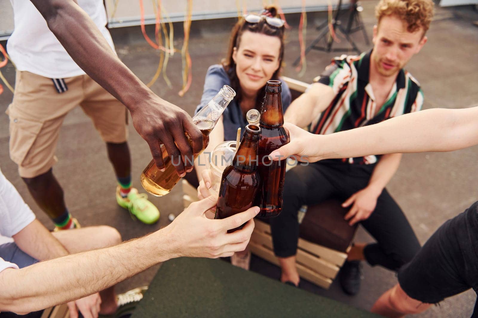 Doing cheers by bottles with beer. Group of young people in casual clothes have a party at rooftop together at daytime.