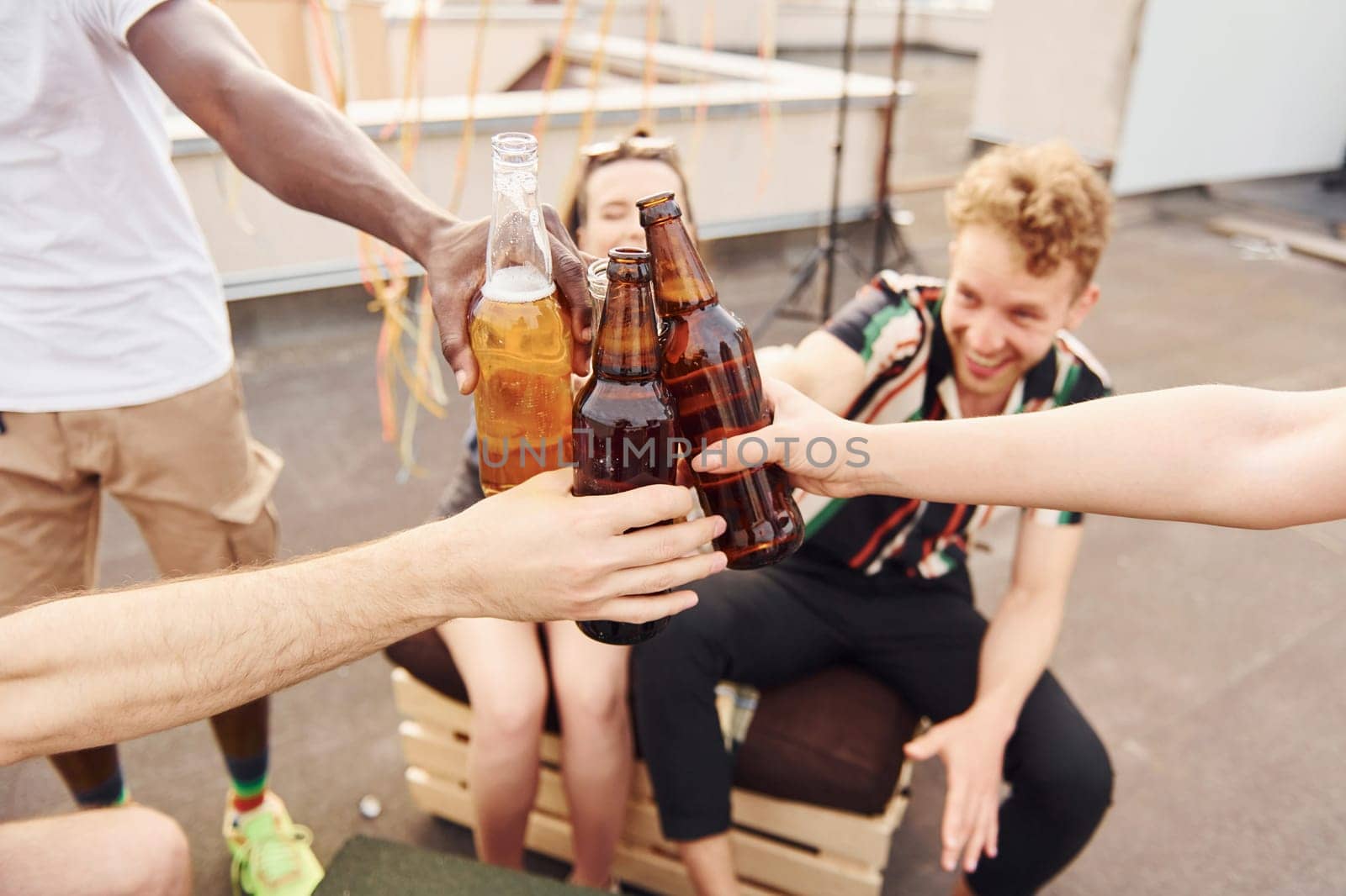 Doing cheers by bottles with beer. Group of young people in casual clothes have a party at rooftop together at daytime.