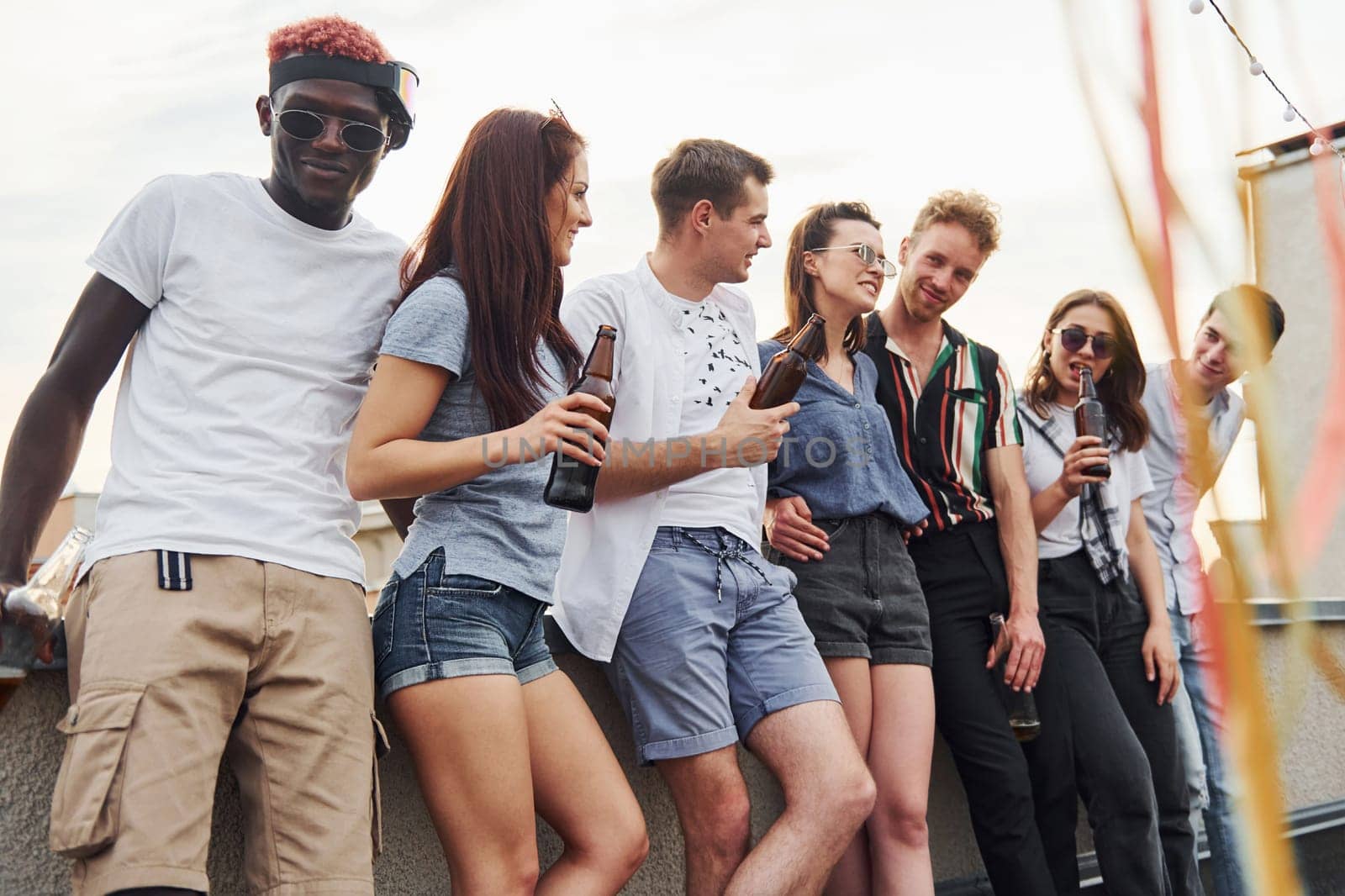 Leaning on the edge of the rooftop with decorates. Group of young people in casual clothes have a party together at daytime.