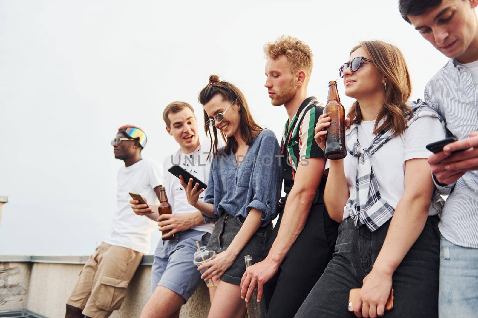 Standing with phones and alcohol in hands. Group of young people in casual clothes have a party at rooftop together at daytime.