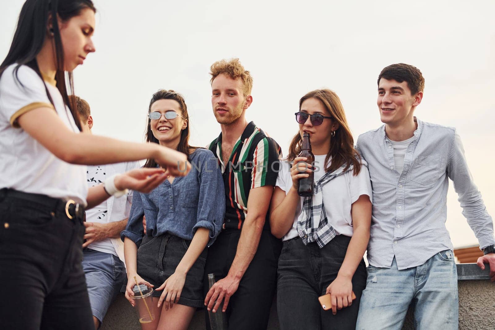 Standing with phones and alcohol in hands. Group of young people in casual clothes have a party at rooftop together at daytime.