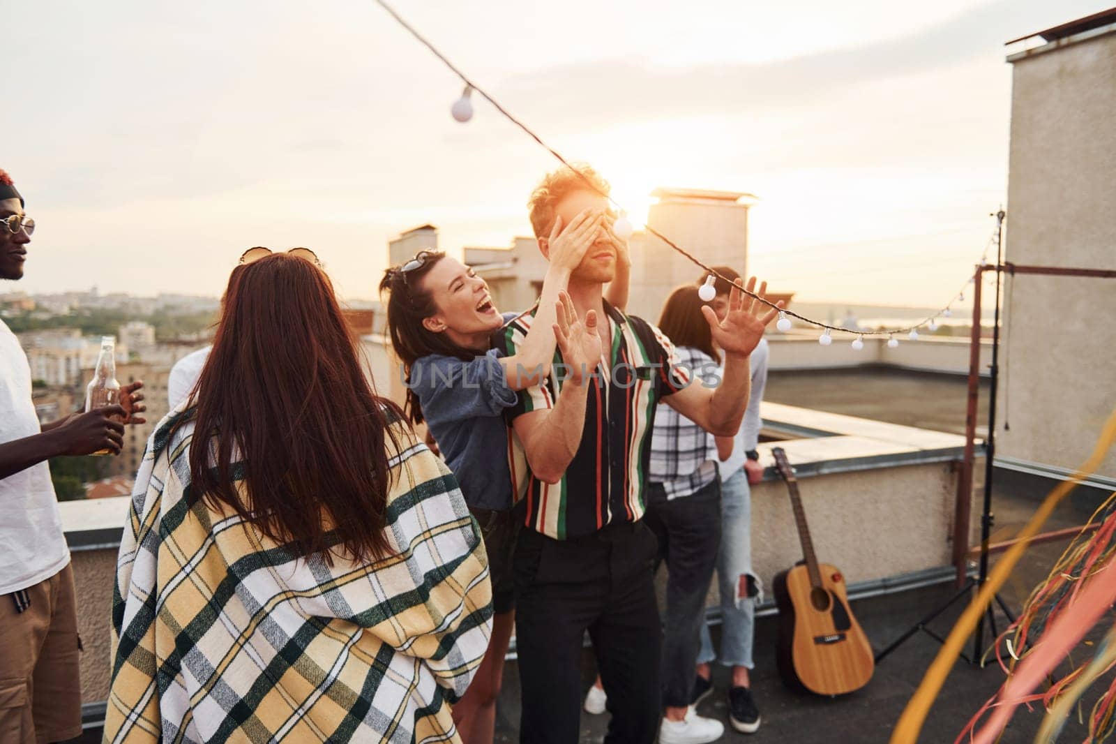 Playing game. Man's eyes covered by hands. Group of young people in casual clothes have a party at rooftop together at daytime.