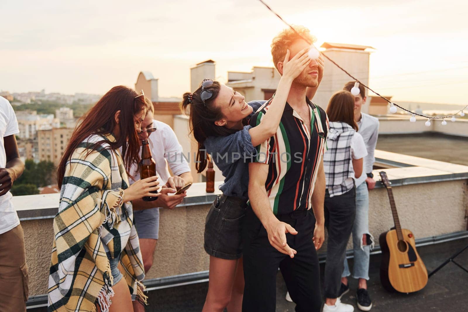 Playing game. Man's eyes covered by hands. Group of young people in casual clothes have a party at rooftop together at daytime.