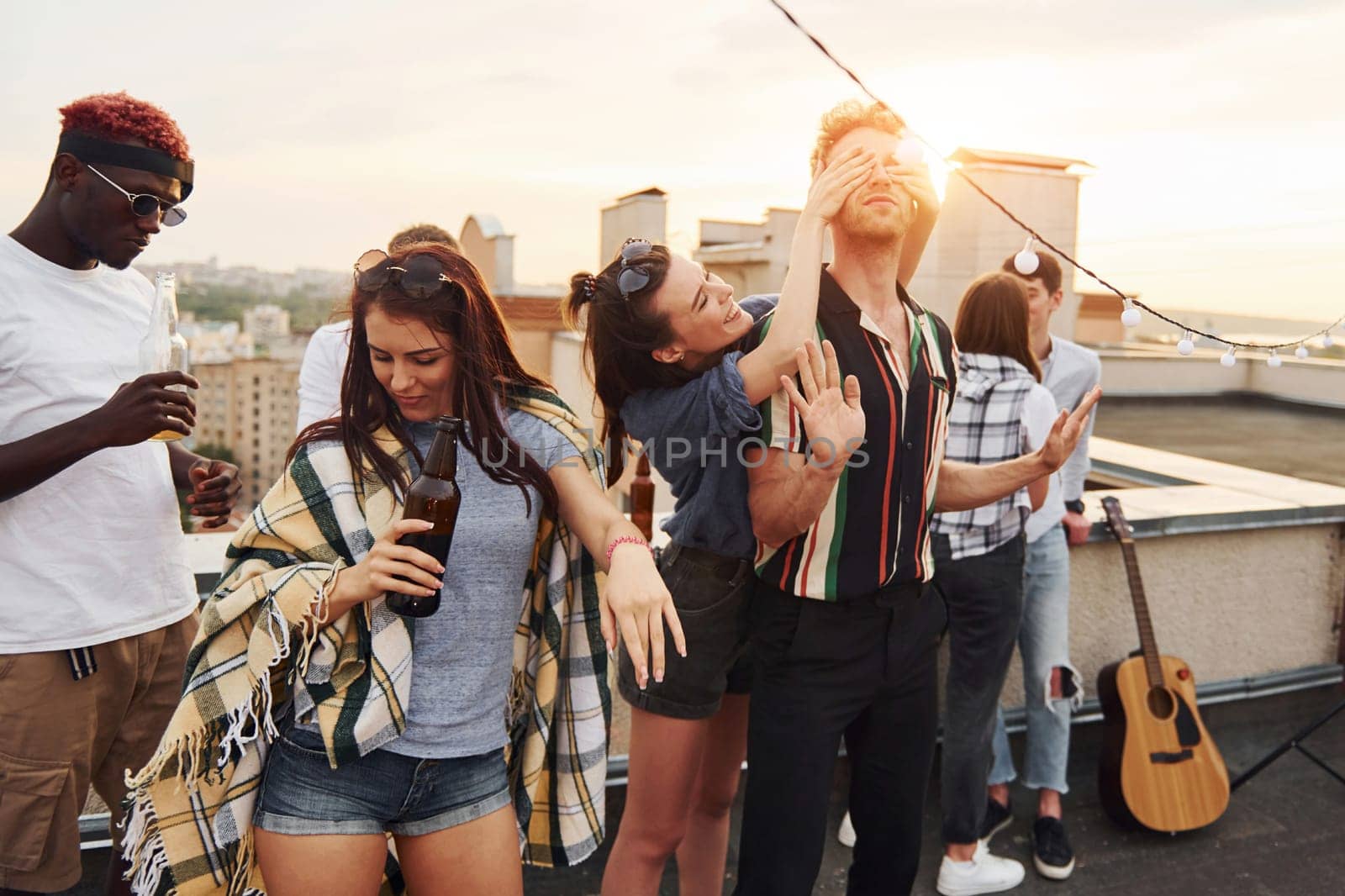 Playing game. Man's eyes covered by hands. Group of young people in casual clothes have a party at rooftop together at daytime.