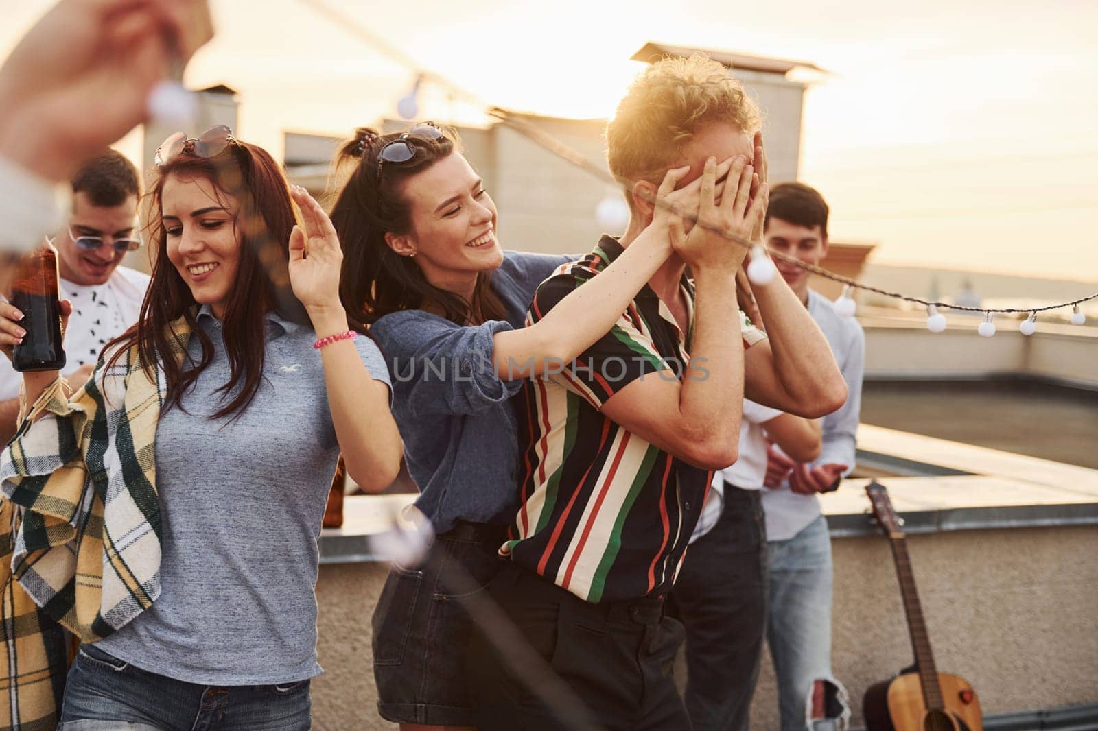 Playing game. Man's eyes covered by hands. Group of young people in casual clothes have a party at rooftop together at daytime.