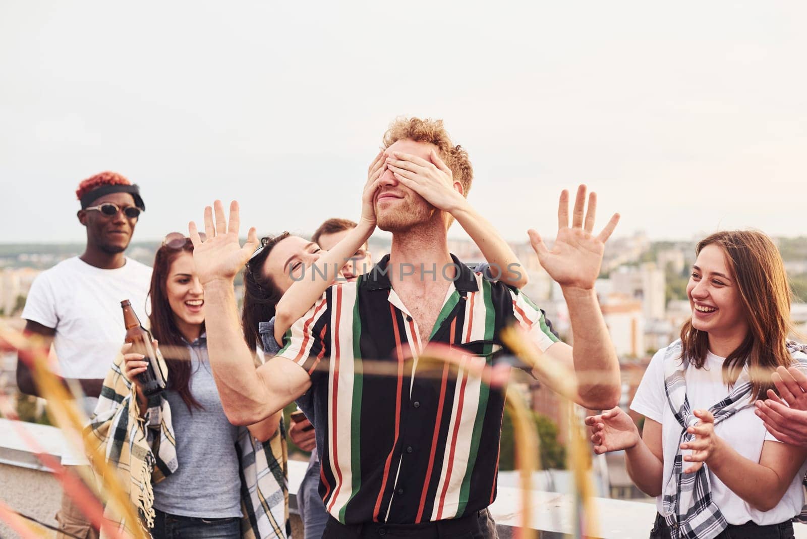 Man's eyes covered by hands. Group of young people in casual clothes have a party at rooftop together at daytime by Standret