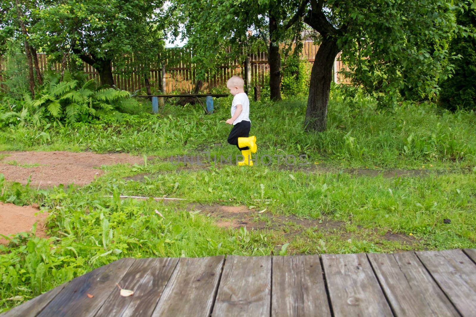 A small, bald-headed boy in yellow boots runs in the countryside through puddles in the fresh air