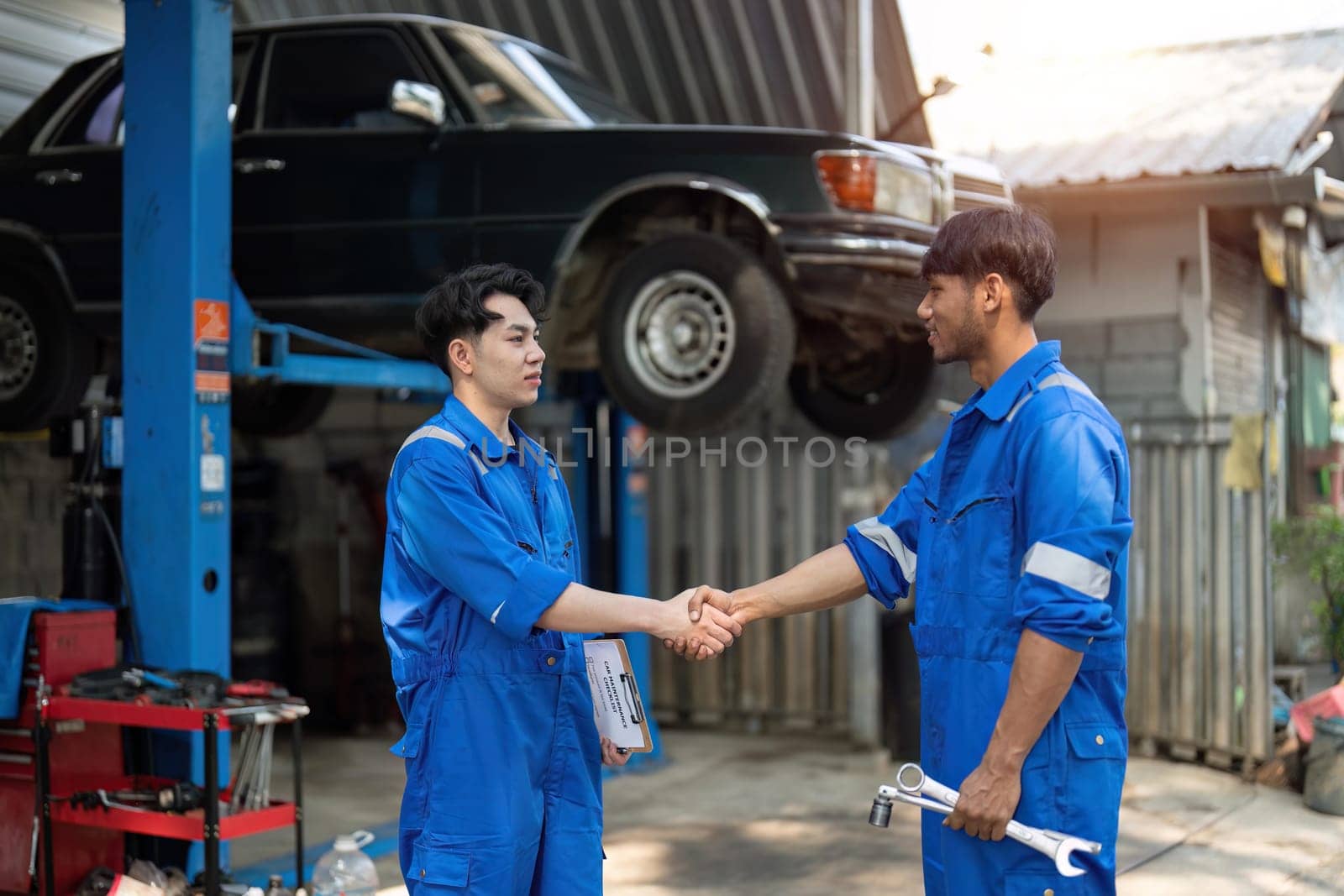 Two Asian automotive mechanic repairman handshake in garage. Vehicle service manager working in mechanics workshop, feel happy and success after check and maintenance car engine for customer. by nateemee