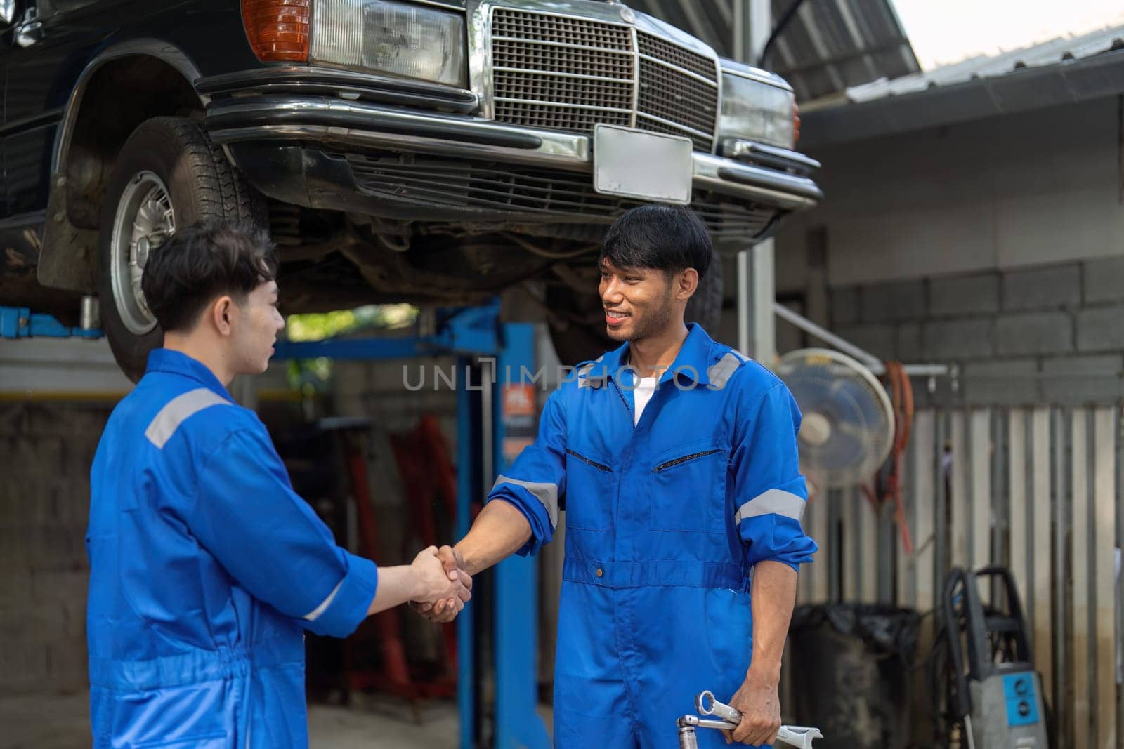 Two Asian automotive mechanic repairman handshake in garage. Vehicle service manager working in mechanics workshop, feel happy and success after check and maintenance car engine for customer. by nateemee