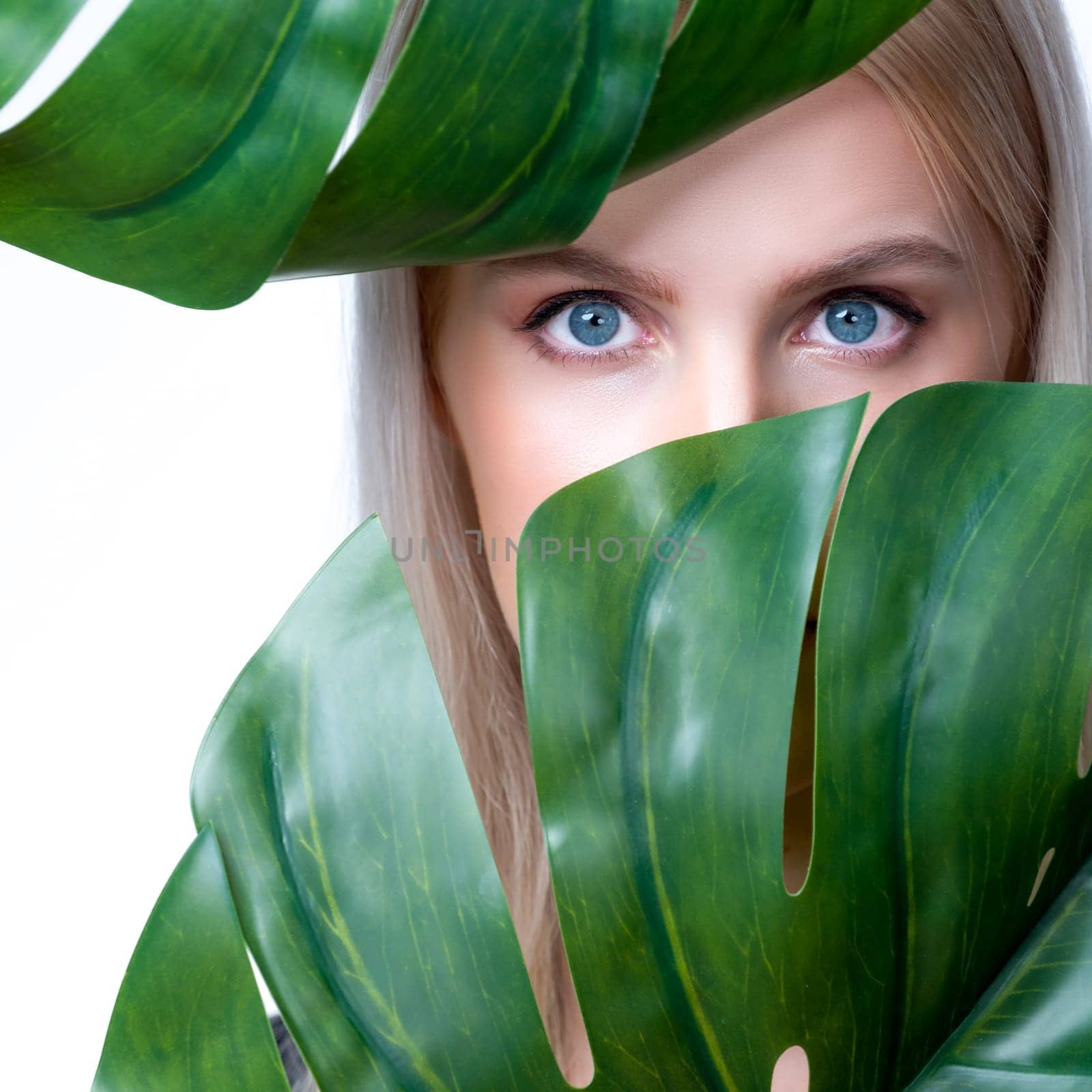 Closeup facial portrait personable woman with perfect smooth makeup holding green monstera leaves and cover her face as natural healthy skincare treatment. Tropical nature and beauty concept