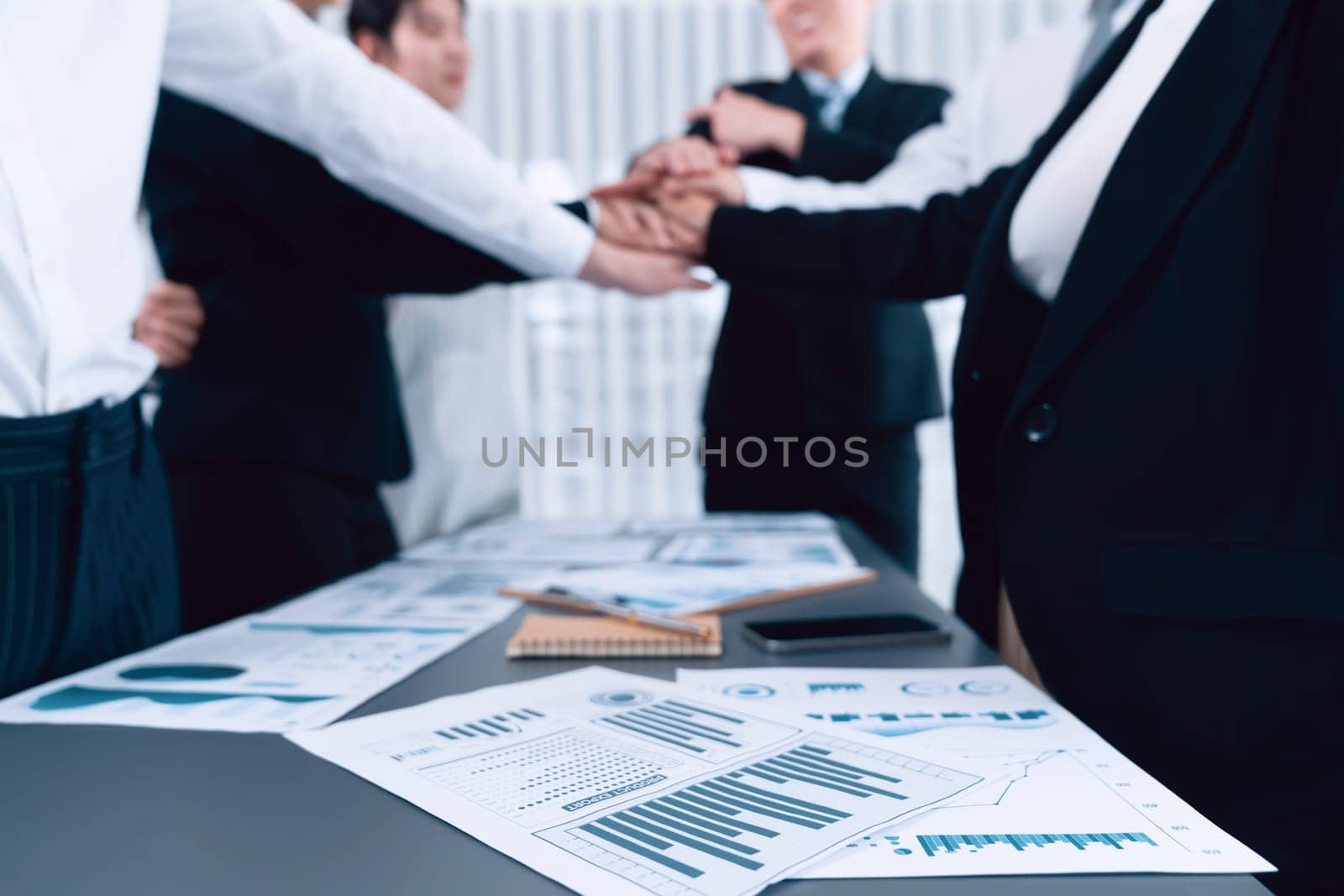 Focus financial report with statistic charts analyzed by BI papers on meeting table at conference room with blur business colleague people put hand stack together in background as harmony concept.