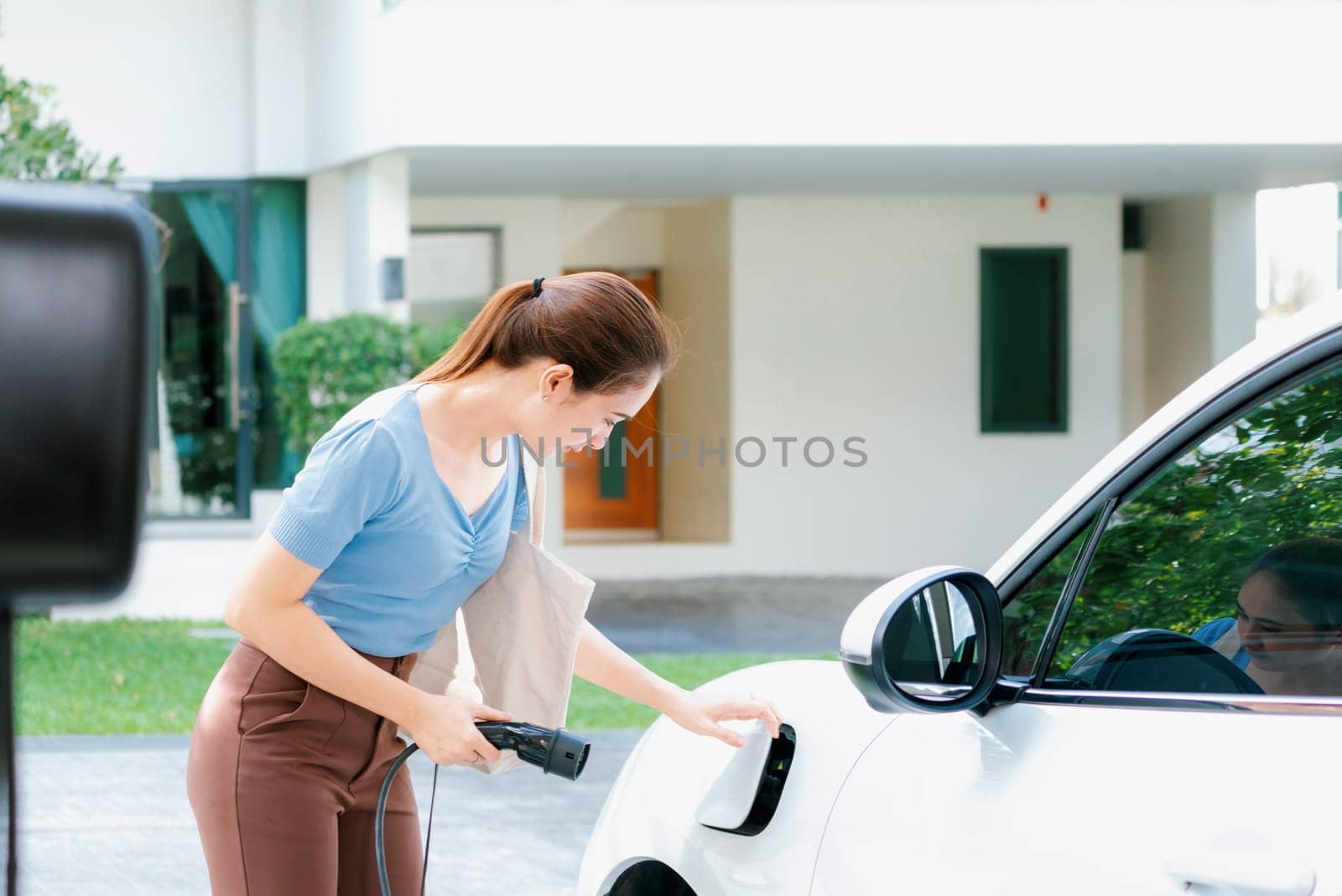 Progressive woman install cable plug to her electric car with home charging station. Concept of the use of electric vehicles in a progressive lifestyle contributes to clean environment.