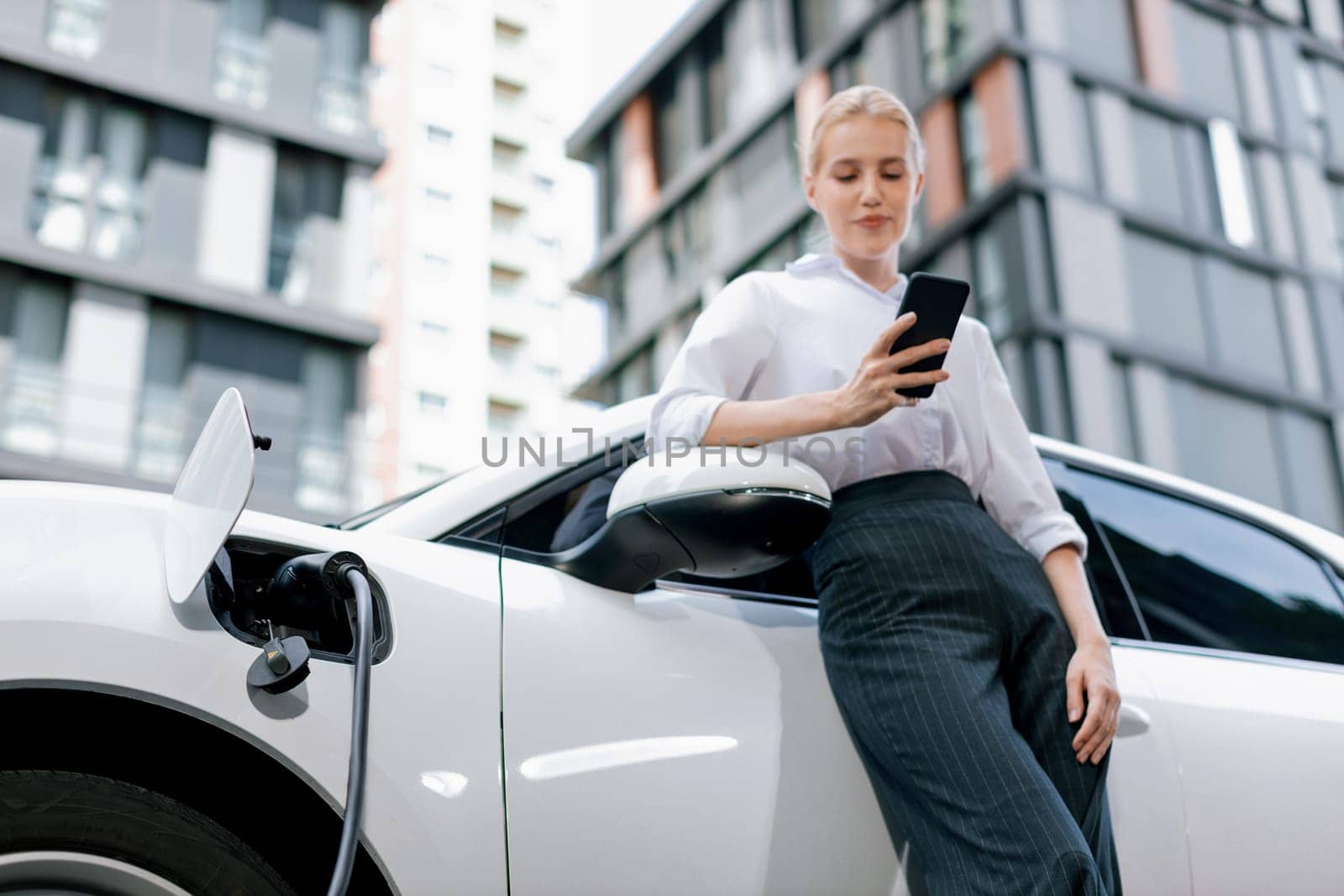 Closeup progressive businesswoman talking on smartphone, leaning electric car at charging station with residential condo apartment in background. Eco friendly rechargeable EV car concept.