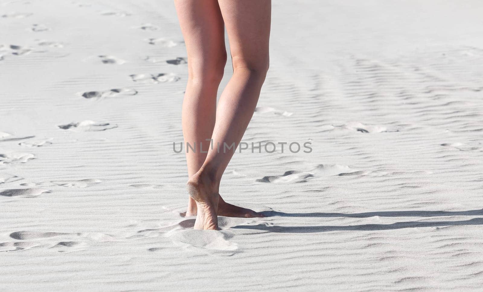 Naked legs of a woman walking on the white sand of the sea on a summer day