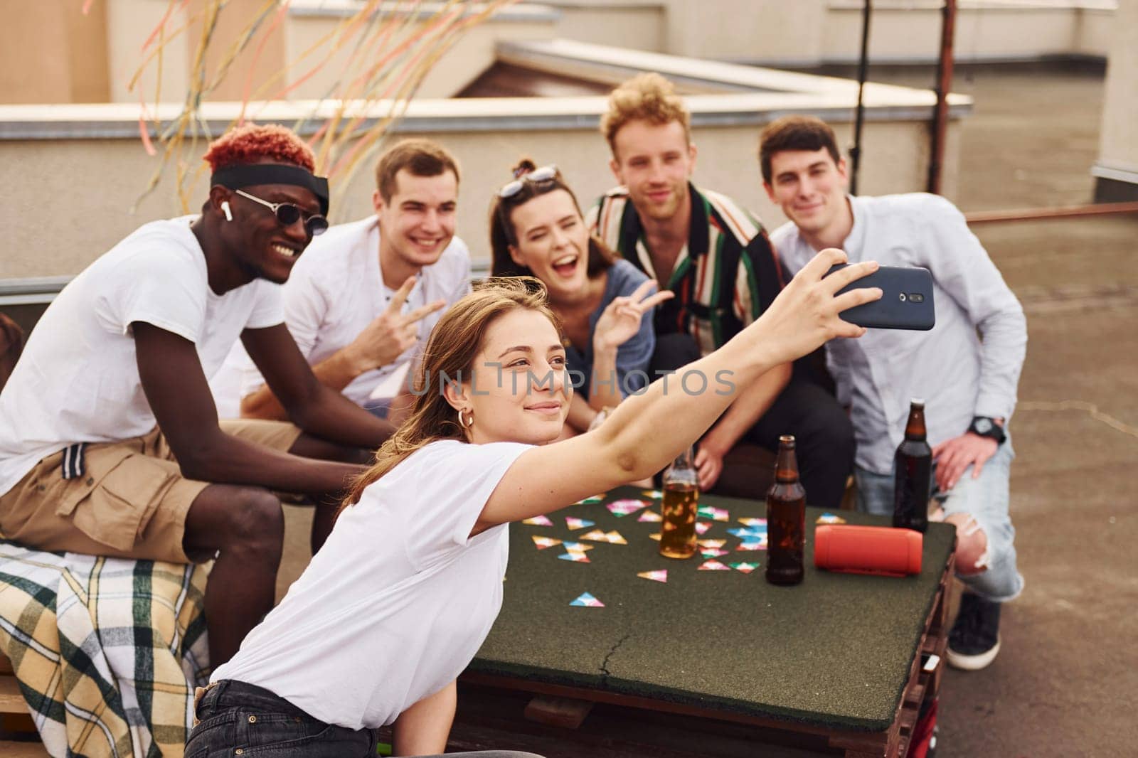 Girl doing photo when people playing card game. Group of young people in casual clothes have a party at rooftop together at daytime.