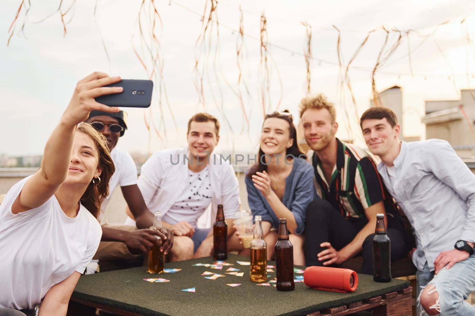 Girl doing photo when people playing card game. Group of young people in casual clothes have a party at rooftop together at daytime.