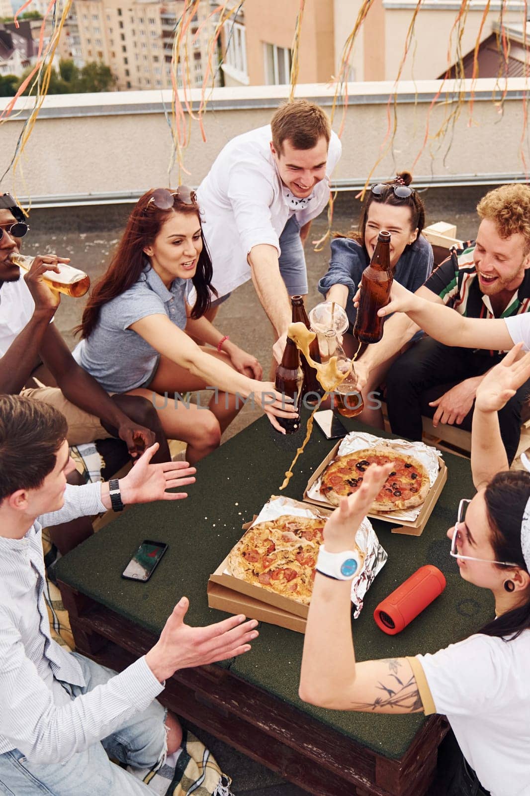 Doing cheers by bottles with beer. Group of young people in casual clothes have a party at rooftop together at daytime by Standret