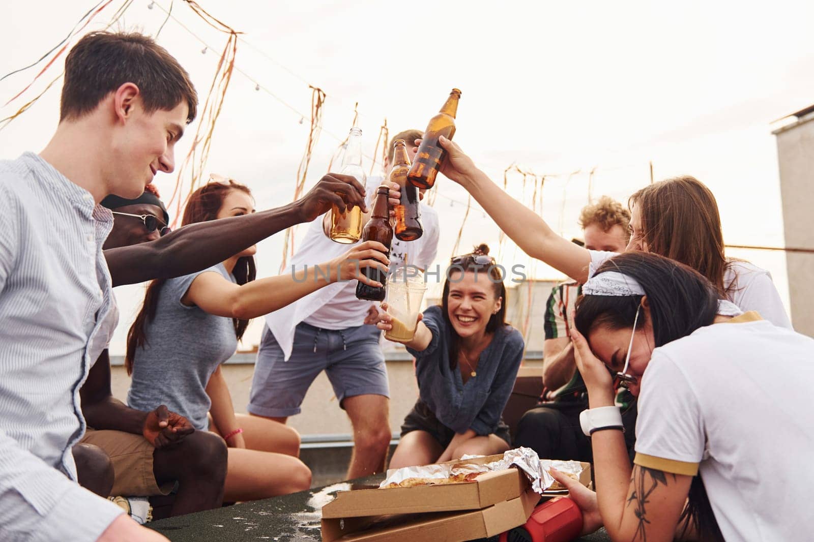 Doing cheers by bottles with beer. Group of young people in casual clothes have a party at rooftop together at daytime.