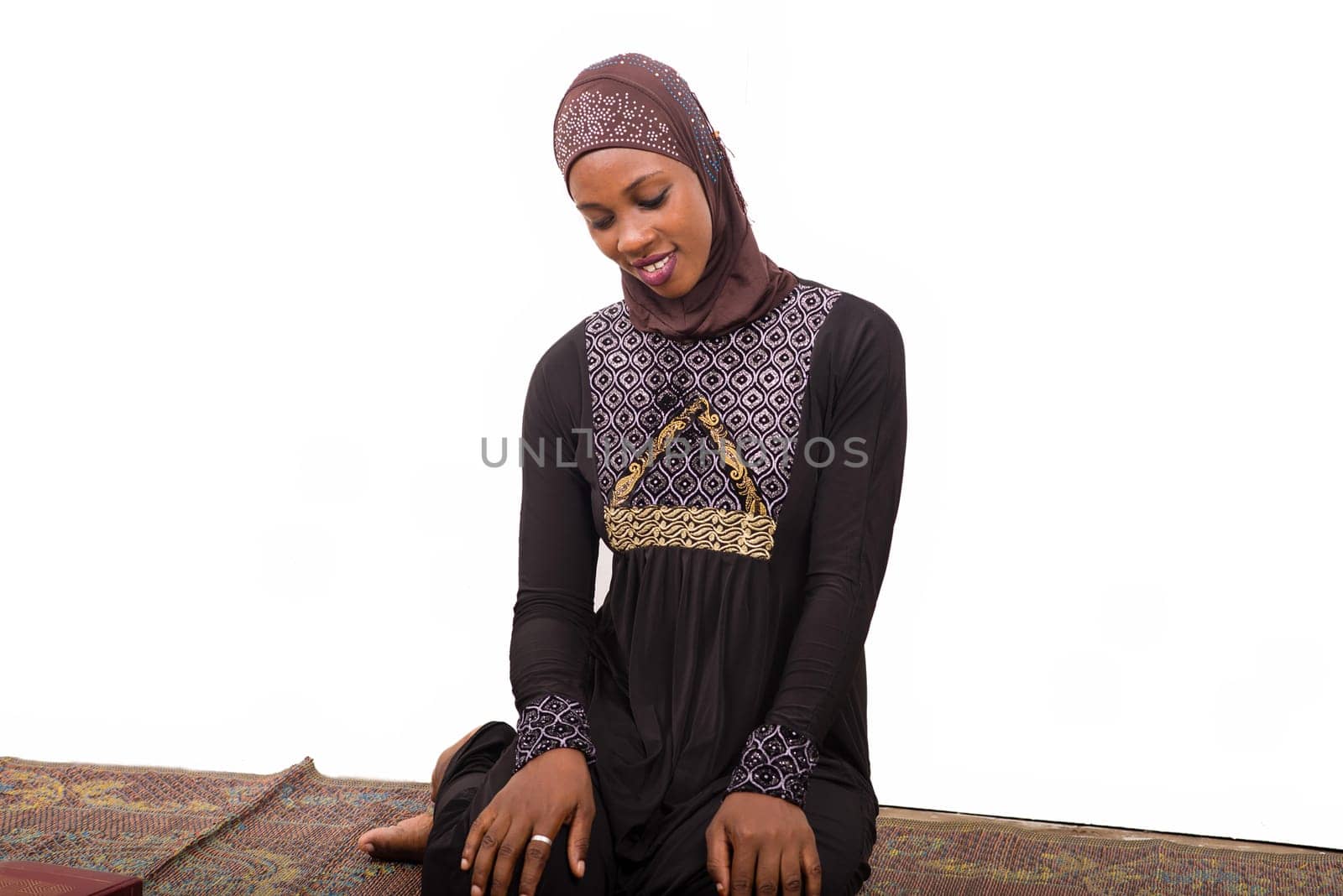 young muslim woman sitting in long dress on mat praying while smiling.