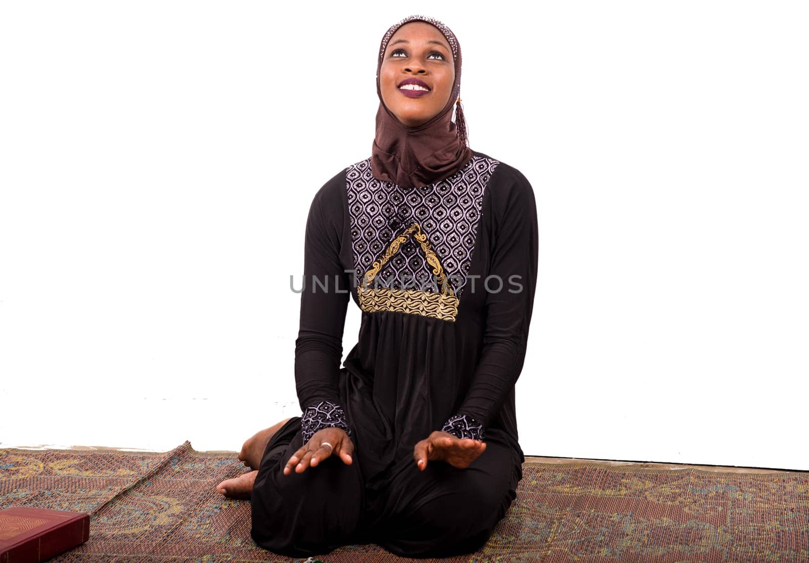 young muslim woman sitting in long dress on mat praying while looking up smiling.