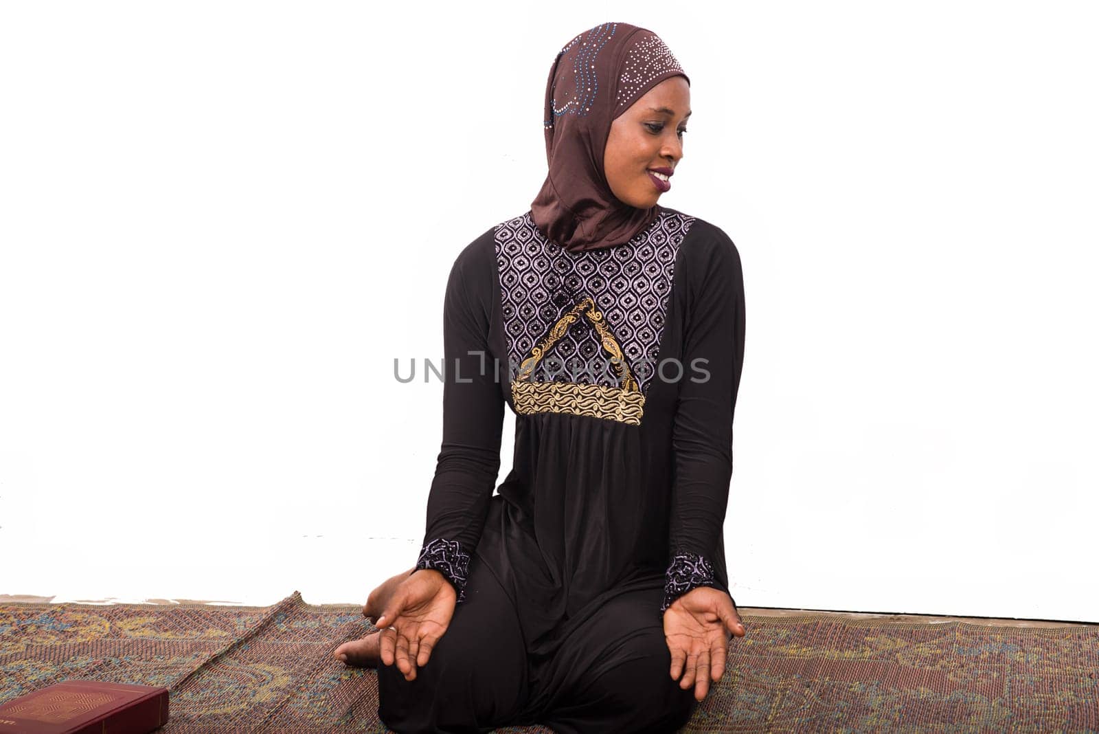 young muslim woman sitting in long dress on mat praying looking at profile smiling.