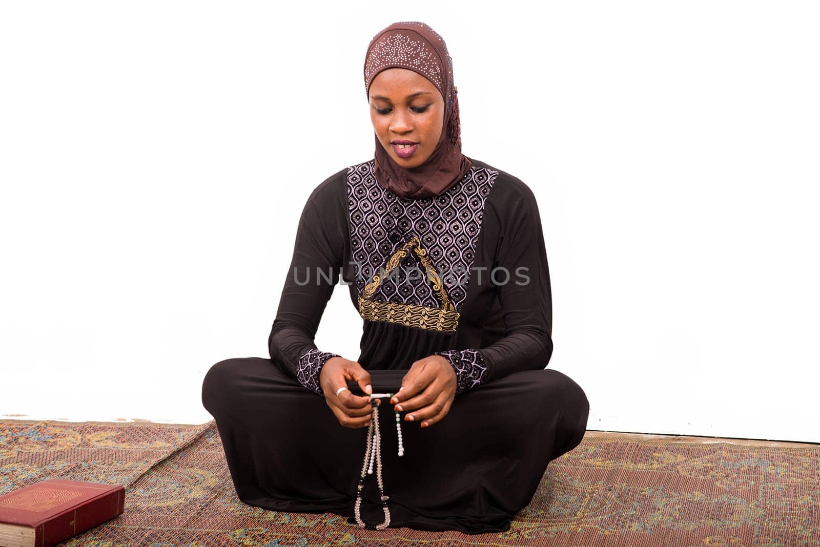 young muslim woman sitting in long dress on mat praying while smiling.