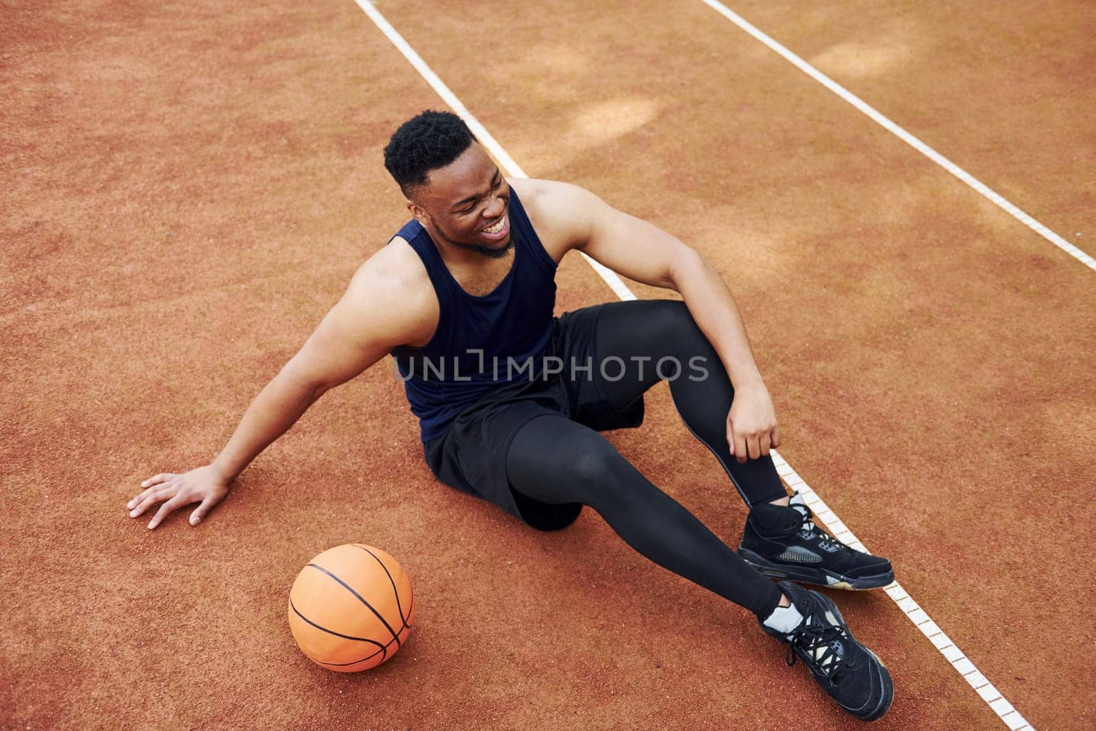 Tired african american man sits on the ground with ball on the court outdoors. Takes a break by Standret