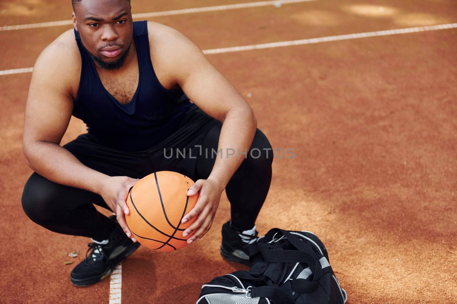 Sits with black bag and preparing for the game. African american man plays basketball on the court outdoors by Standret