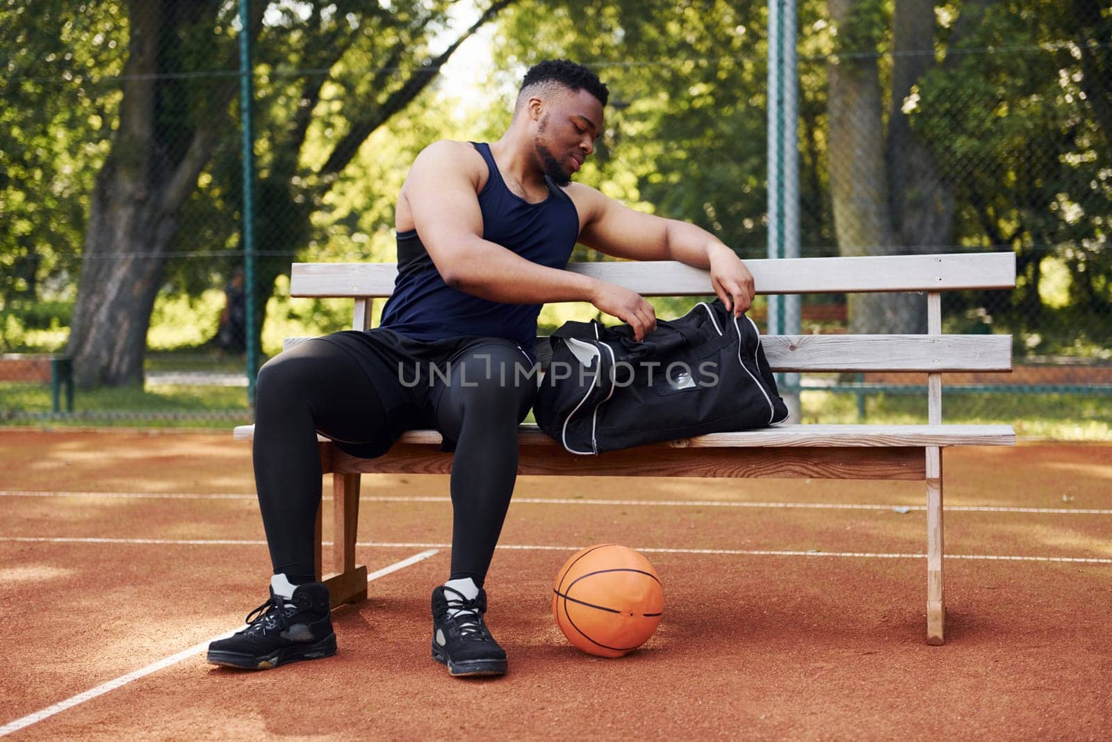 Sits with black bag and preparing for the game. African american man plays basketball on the court outdoors by Standret