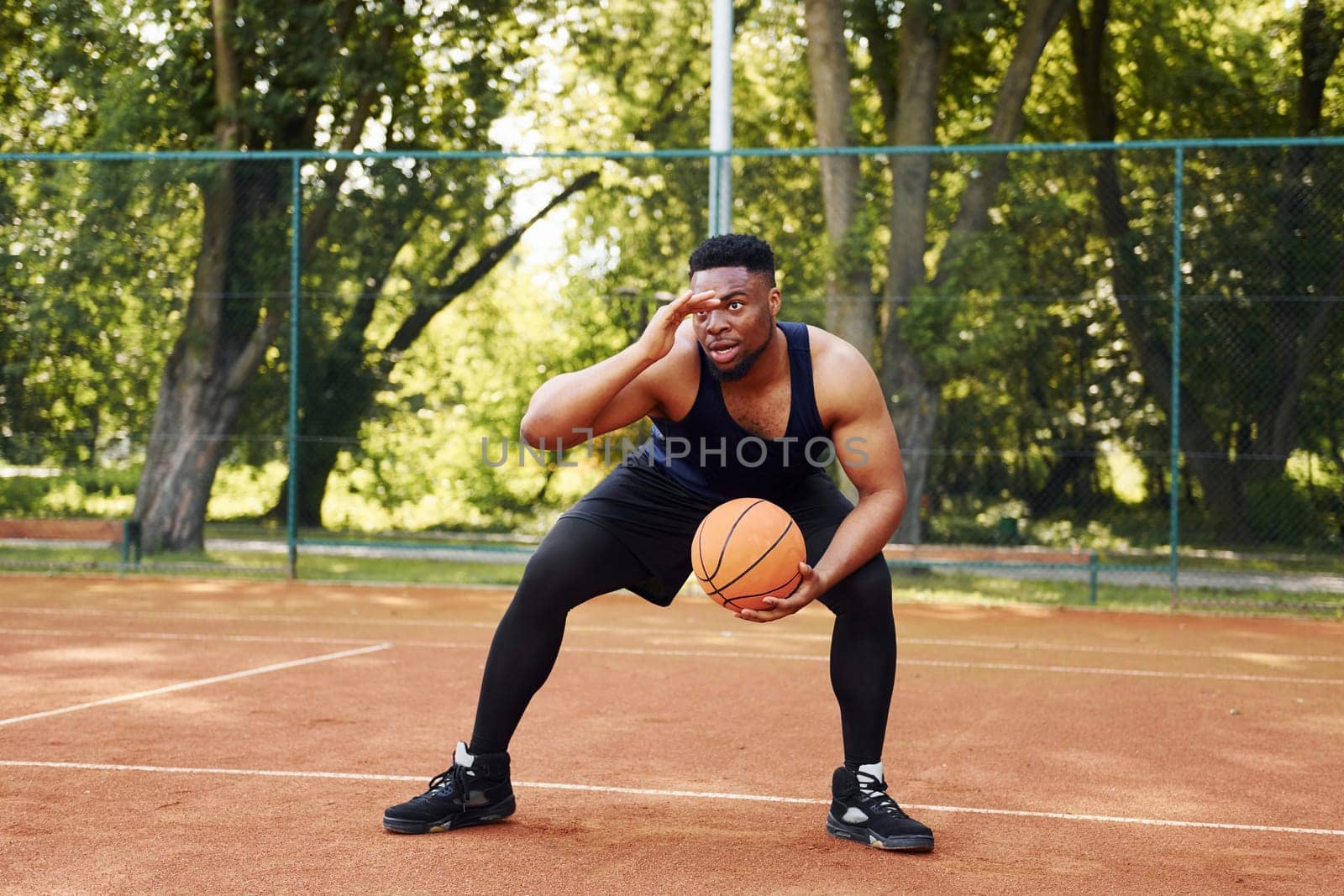 Beautiful green trees on background. African american man plays basketball on the court outdoors.