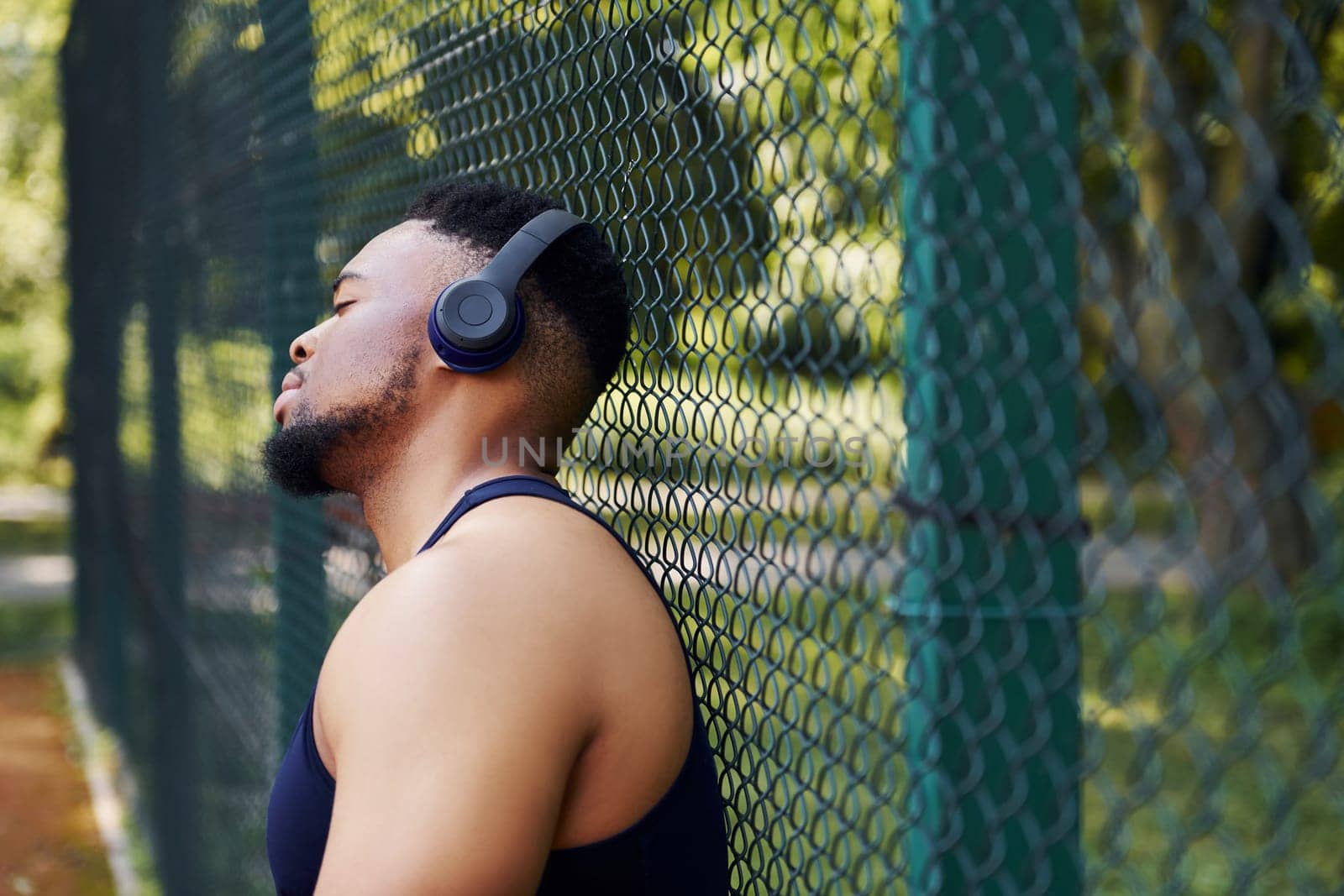 African american guy in wireless headphones leans on the metal mesh of sportive court and takes break by Standret