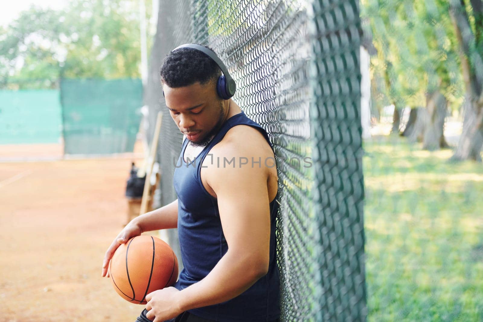 African american man with wireless headphones takes a break and leaning on the metal mesh with ball on the court outdoors.