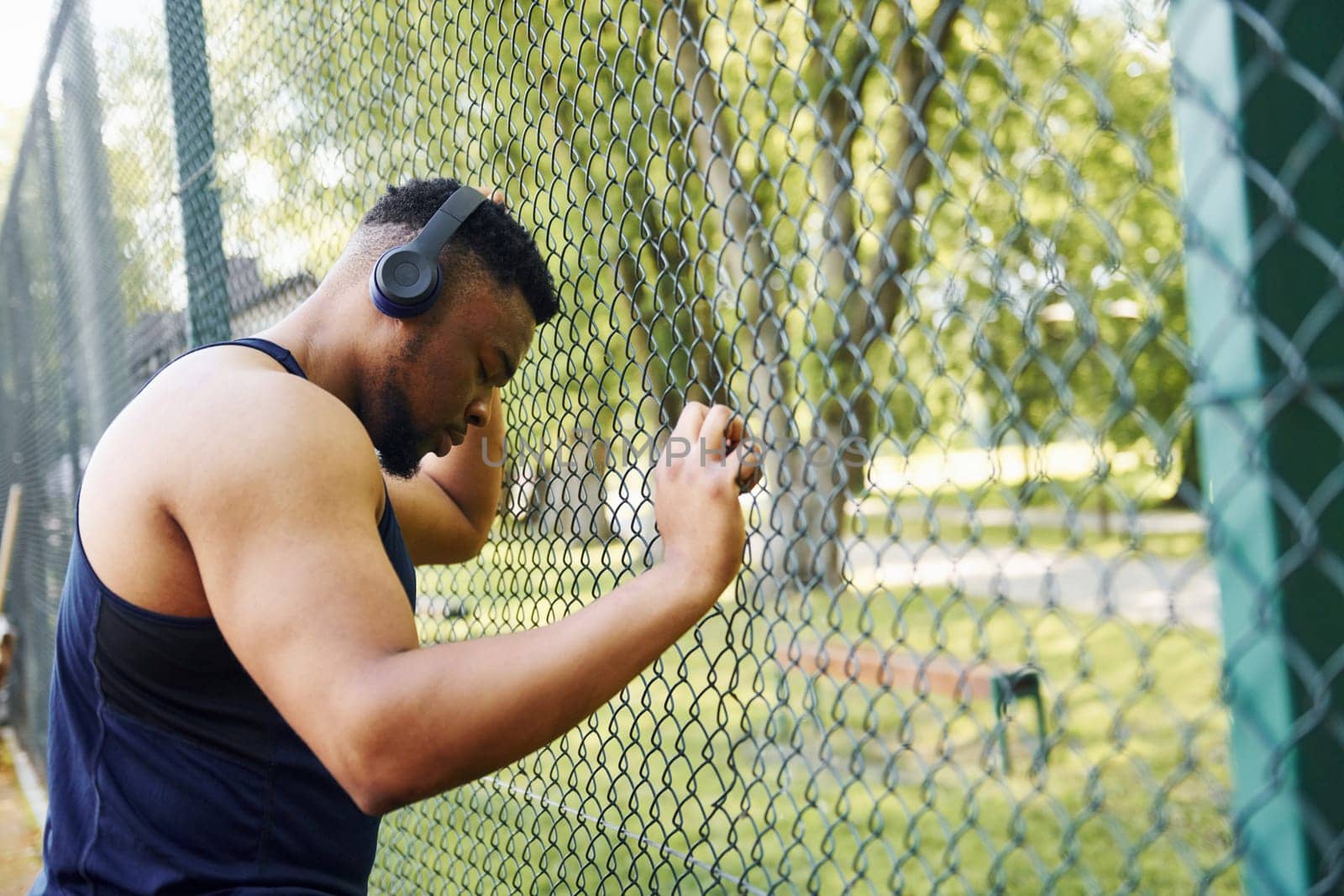 African american guy in wireless headphones leans on the metal mesh of sportive court and takes break.