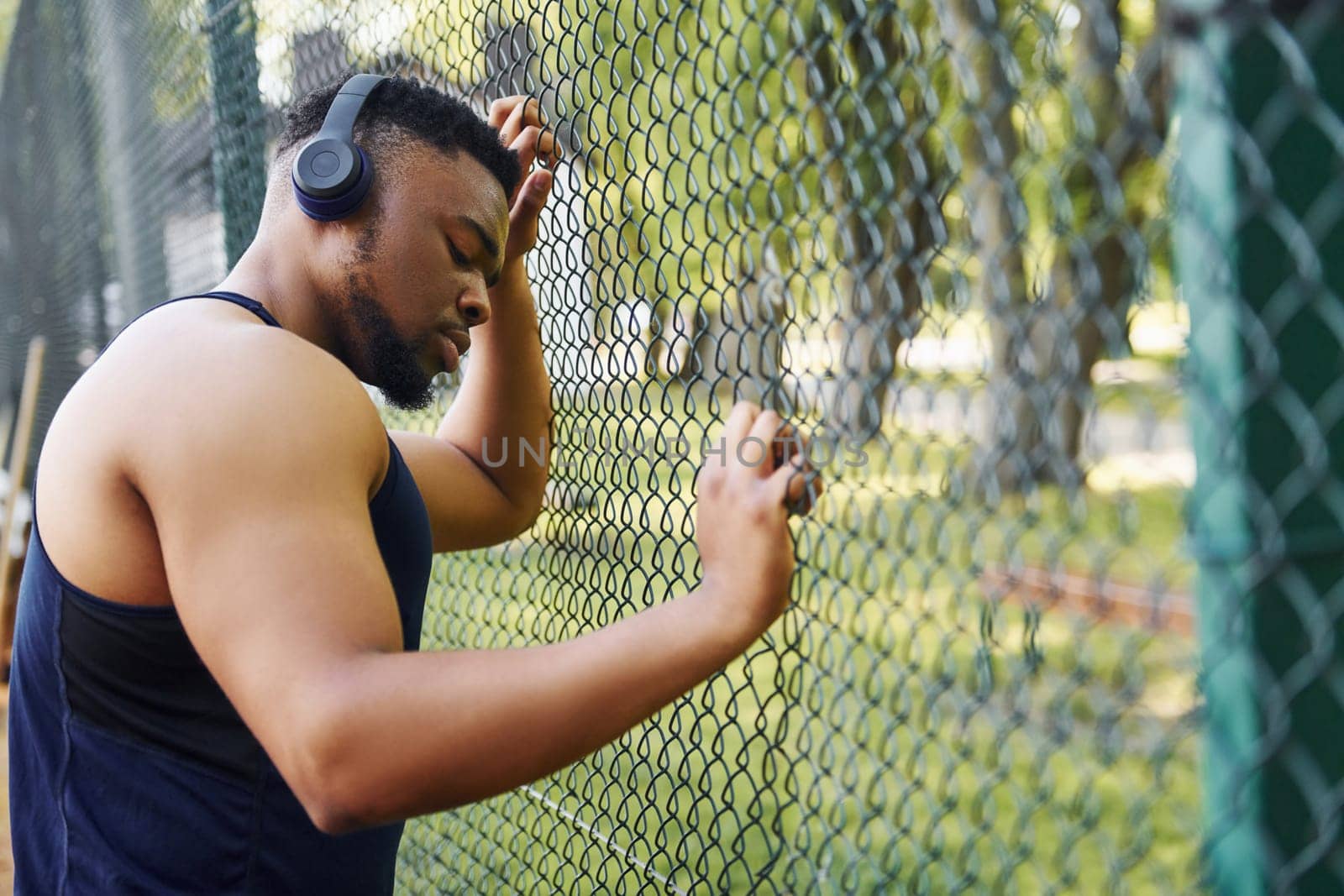 African american guy in wireless headphones leans on the metal mesh of sportive court and takes break.
