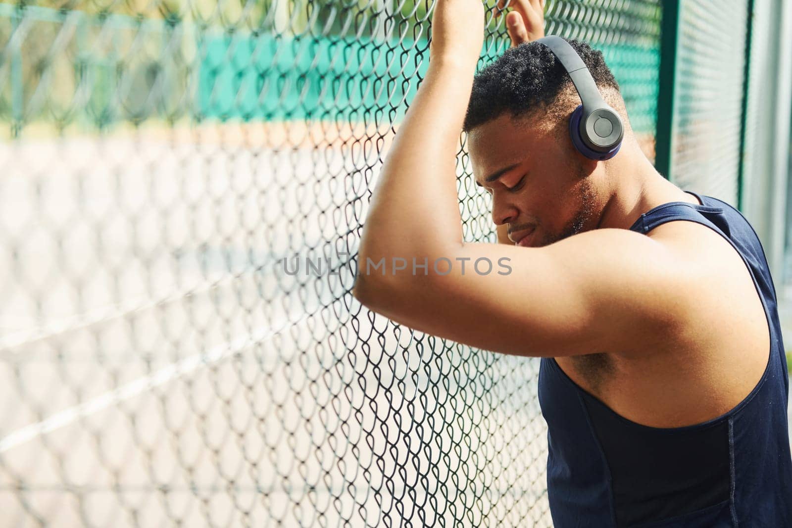 African american guy in wireless headphones leans on the metal mesh of sportive court and takes break by Standret
