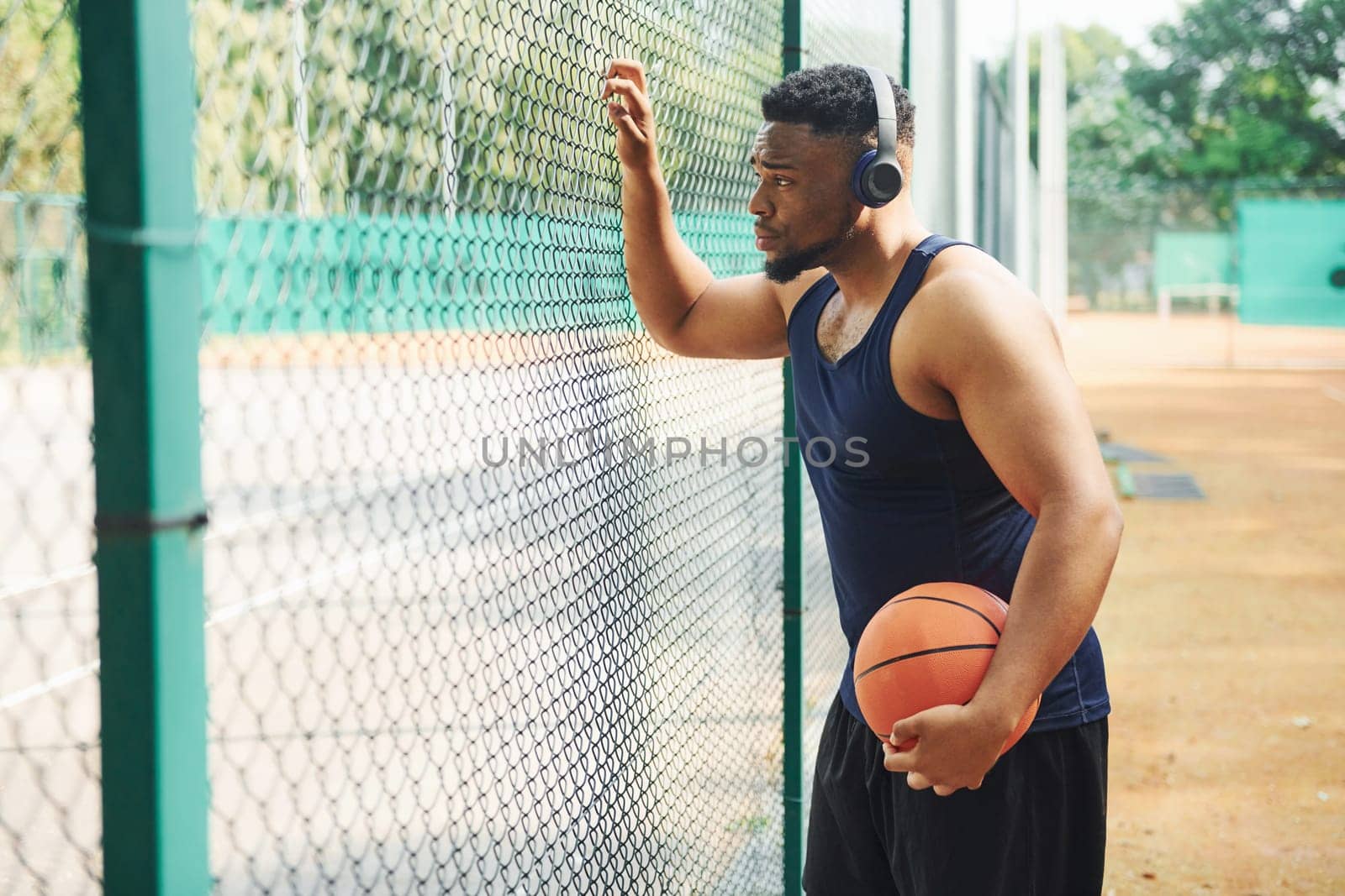 African american man with wireless headphones takes a break and leaning on the metal mesh with ball on the court outdoors by Standret