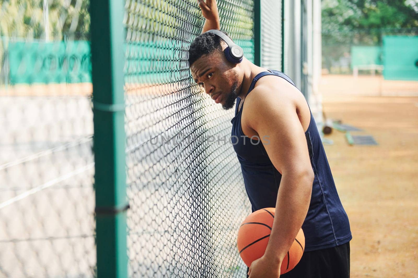 African american man with wireless headphones takes a break and leaning on the metal mesh with ball on the court outdoors by Standret