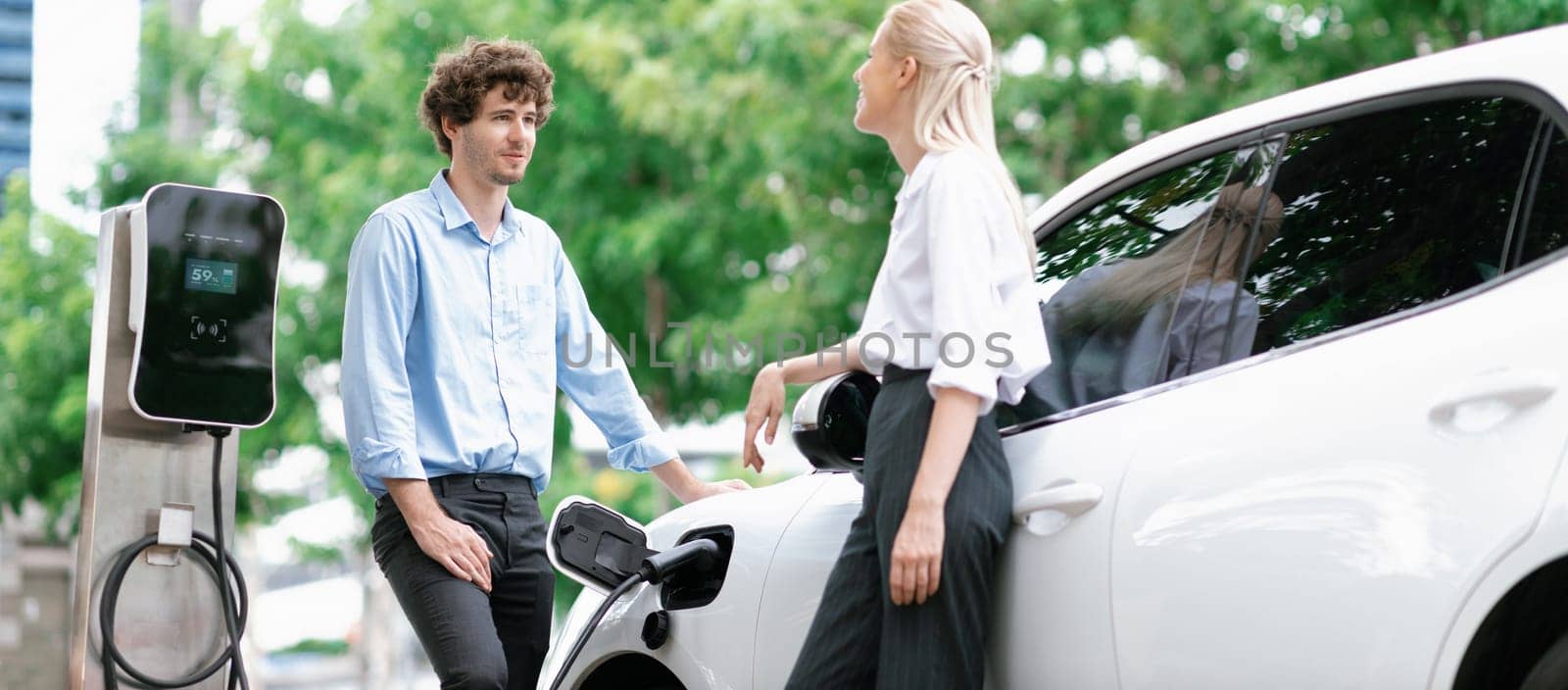 Progressive businessman and businesswoman with electric car parking and connected to public charging station before driving around city center. Eco friendly rechargeable car powered by clean energy.