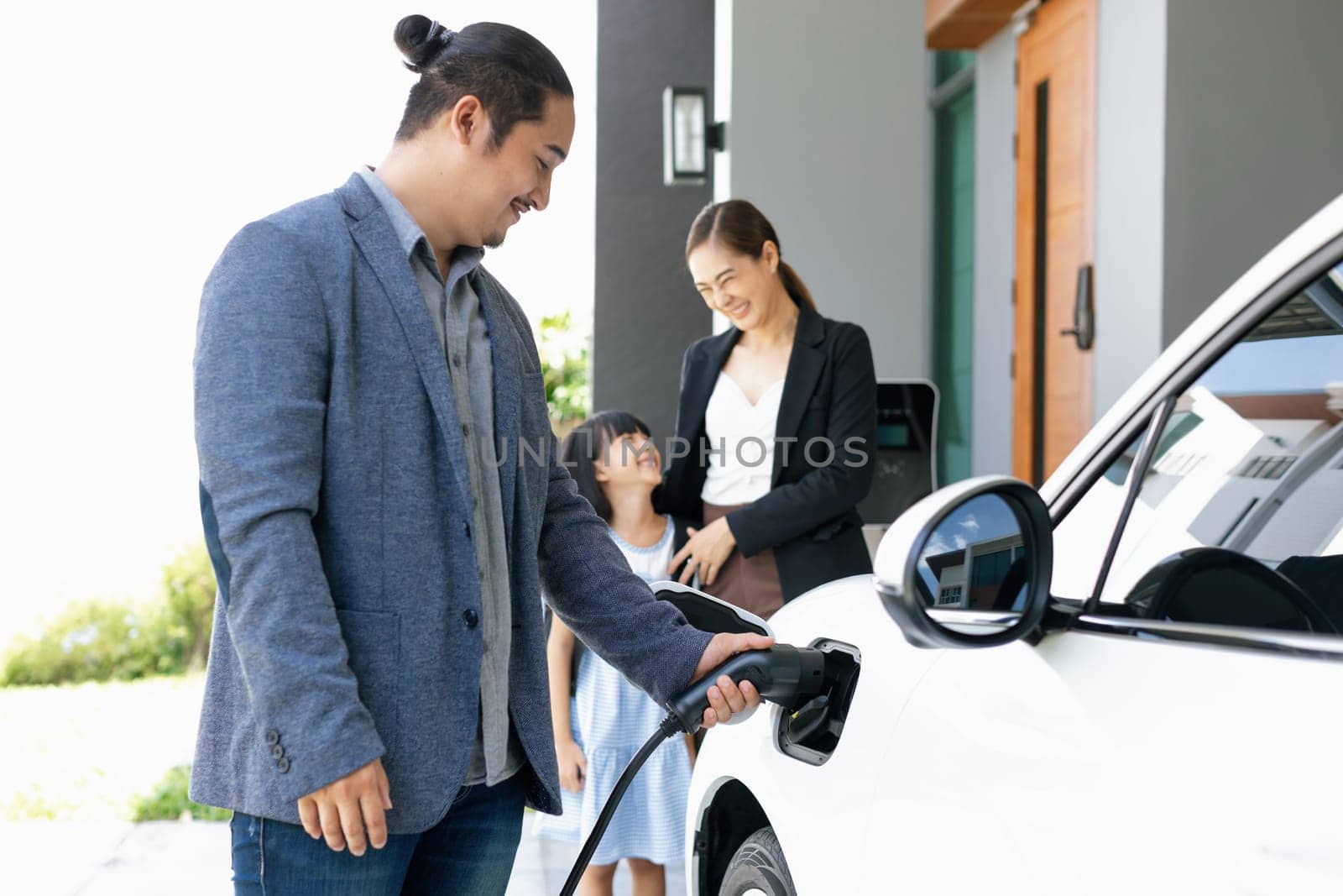 Progressive young parents and daughter with electric vehicle and home charging station. Green and clean energy from electric vehicles for healthy environment. Eco power from renewable source at home.