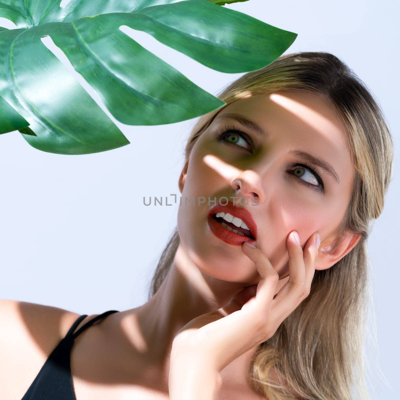 Closeup young blond hair woman with perfect skin and soft alluring facial makeup cover face with green leaf monstera from bright sunlight in isolated background for skincare sunscreen product.
