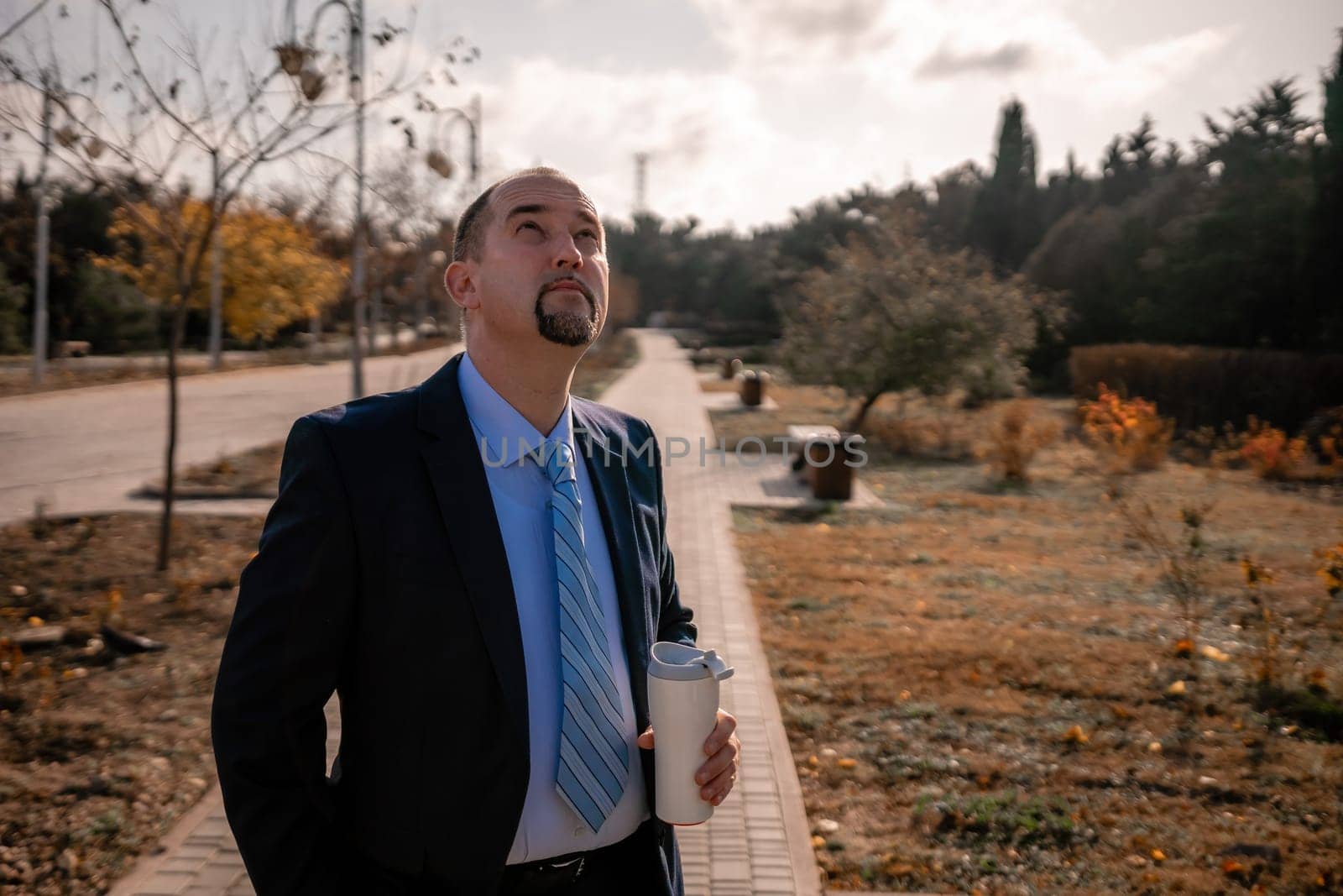 Mature entrepreneur drink coffee in autumn park. Senior executive rest in city park. by panophotograph