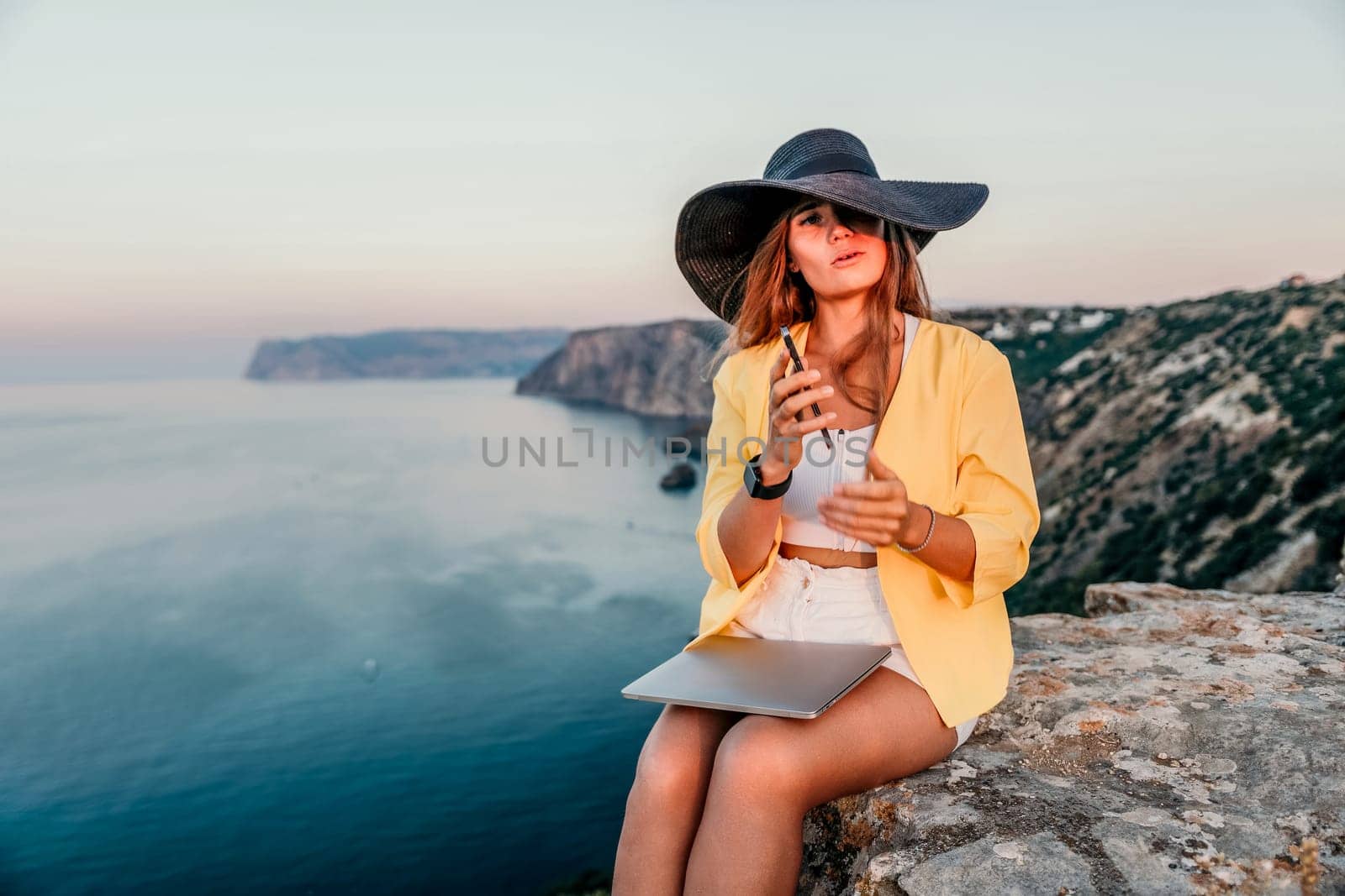 Successful business woman in yellow hat working on laptop by the sea. Pretty lady typing on computer at summer day outdoors. Freelance, travel and holidays concept.