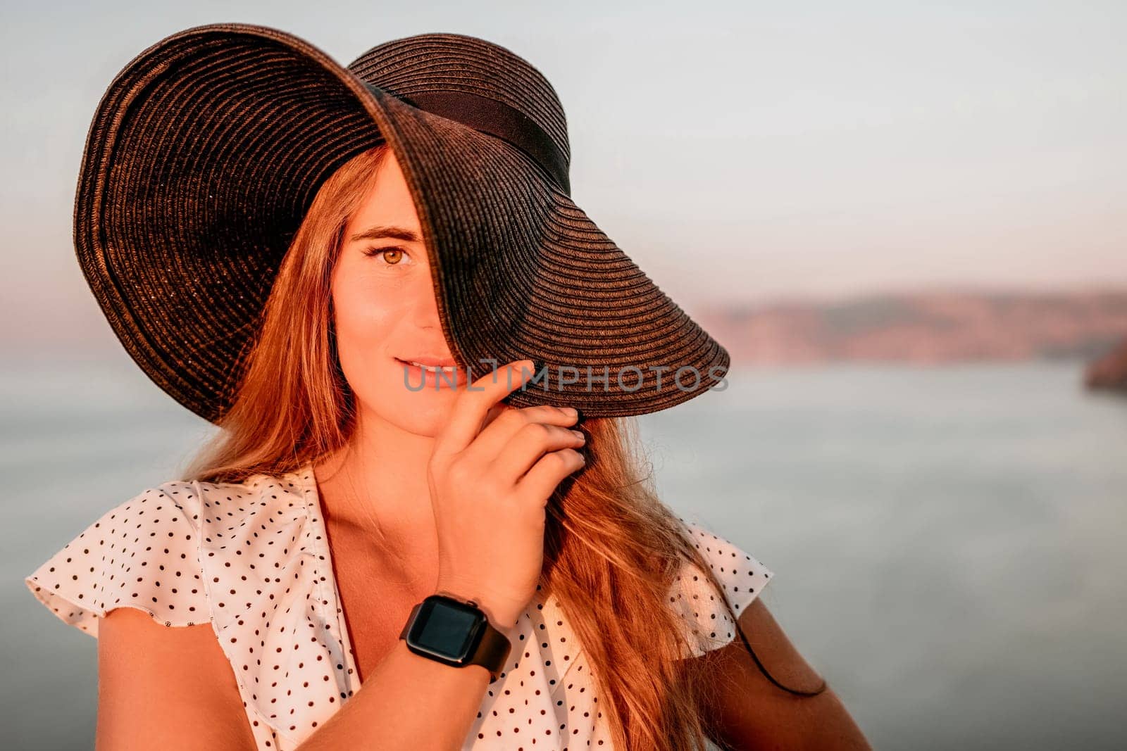 Portrait of happy young woman wearing summer black hat with large brim at beach on sunset. Closeup face of attractive girl with black straw hat. Happy young woman smiling and looking at camera at sea