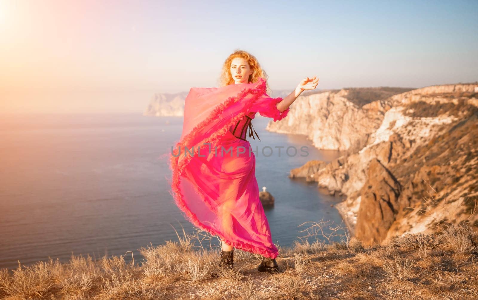 Side view a Young beautiful sensual woman in a red long dress posing on a volcanic rock high above the sea during sunset. Girl on the nature on blue sky background. Fashion photo