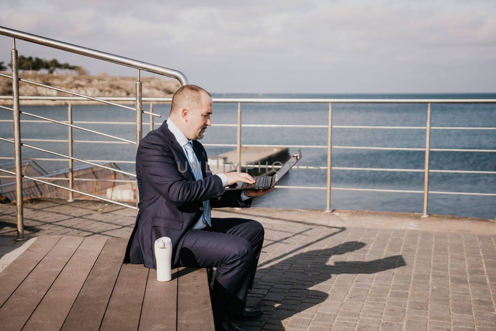 Confident middle age businessman working remotely online, typing on a laptop keyboard while sitting on a beach at sunset. Working remotely on vacation, running an online business from a distance. by panophotograph