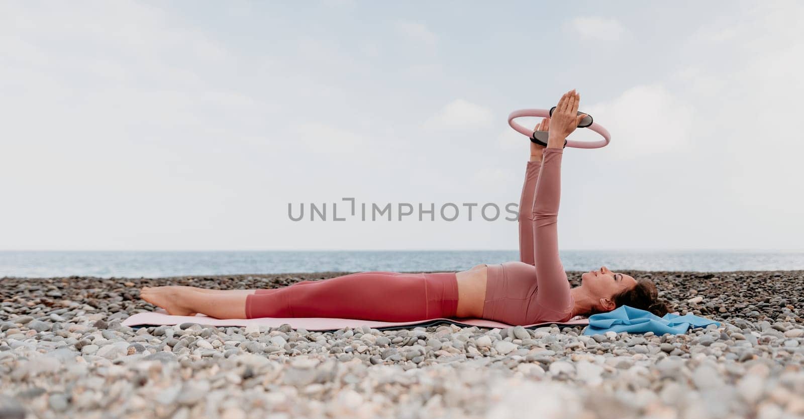 Middle aged well looking woman with black hair doing Pilates with the ring on the yoga mat near the sea on the pebble beach. Female fitness yoga concept. Healthy lifestyle, harmony and meditation.