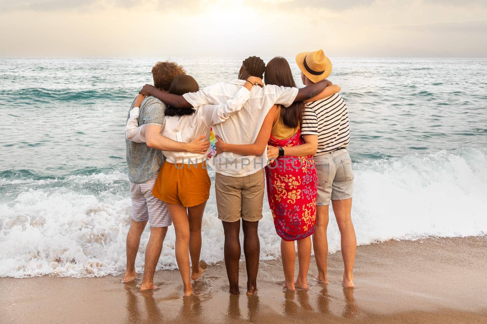 Rear view of multiracial friends embracing together looking at the ocean relaxing during vacation trip. Friendship concept.